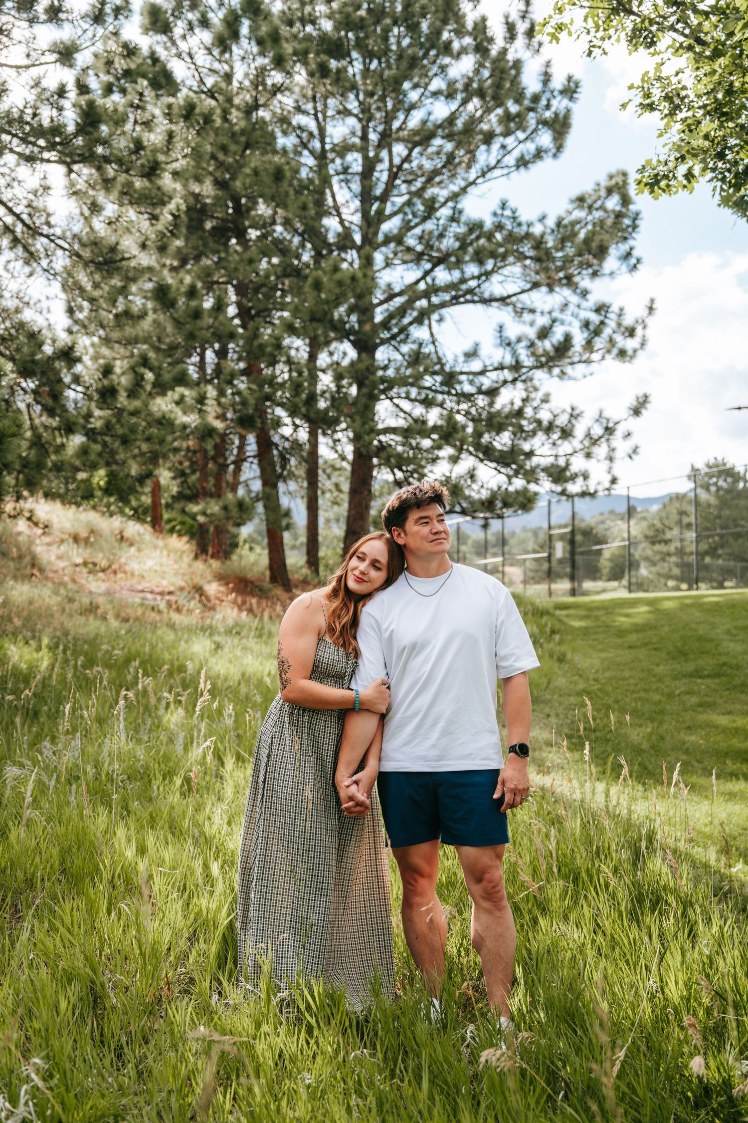 A couple holds hands in a lush green meadow surrounded by pine trees, the woman in a plaid dress leaning on the man.