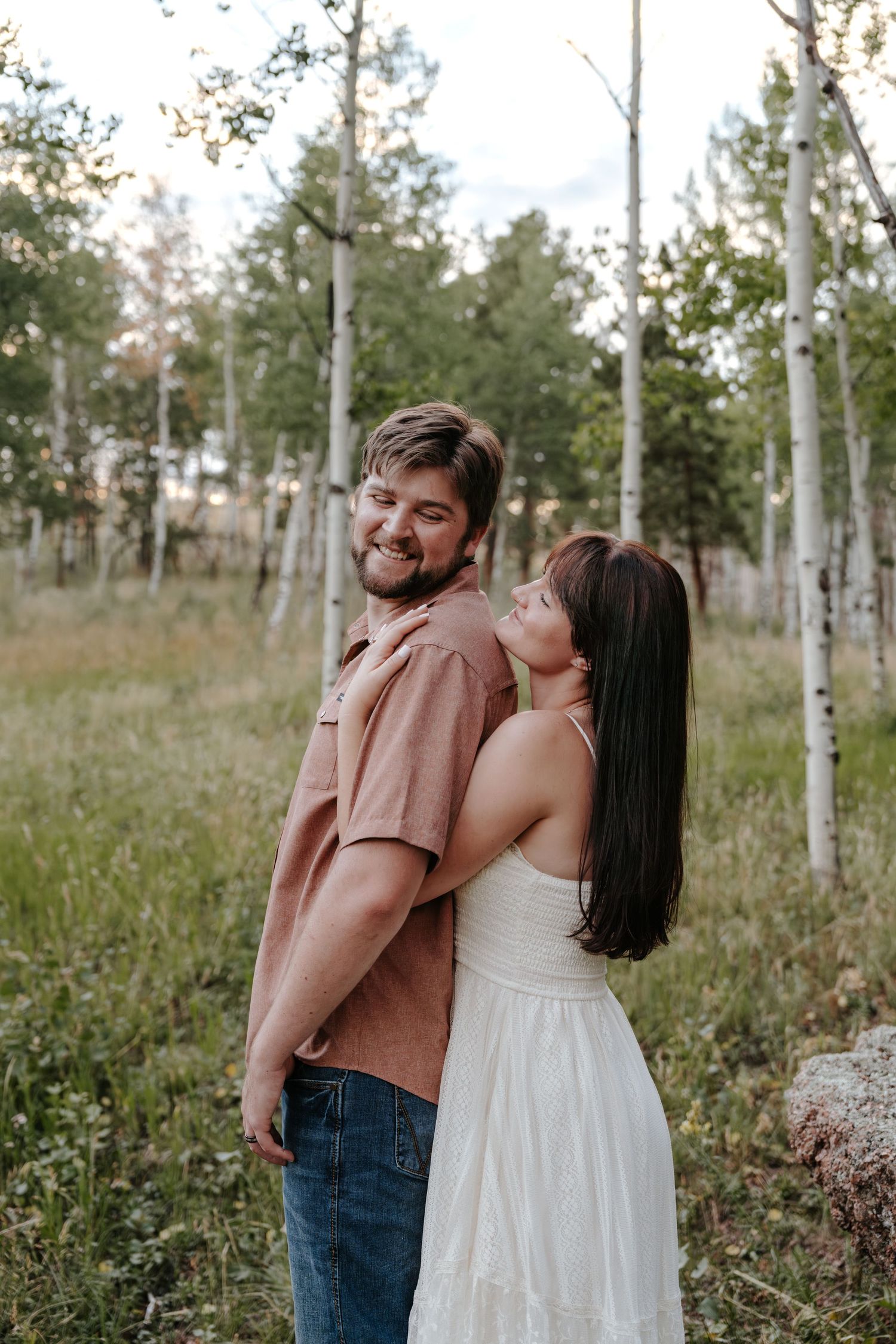 A smiling couple poses in an aspen forest, the woman in a white dress leaning on the man's shoulder.