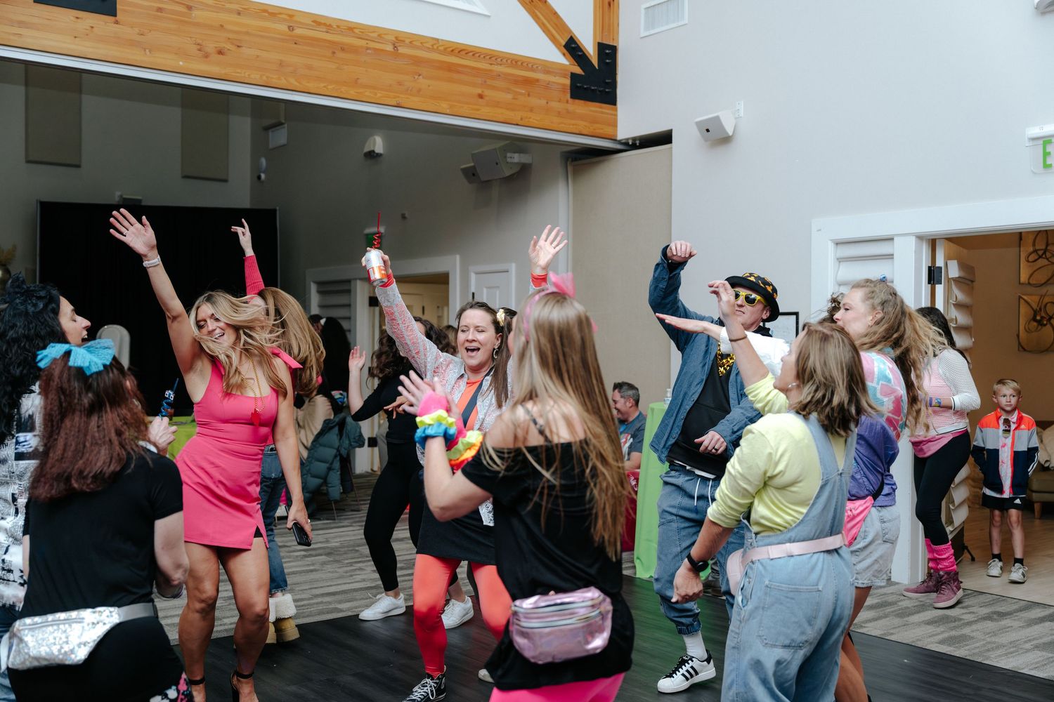 Group of people dancing and celebrating in colorful 80s-themed outfits at an indoor party event.