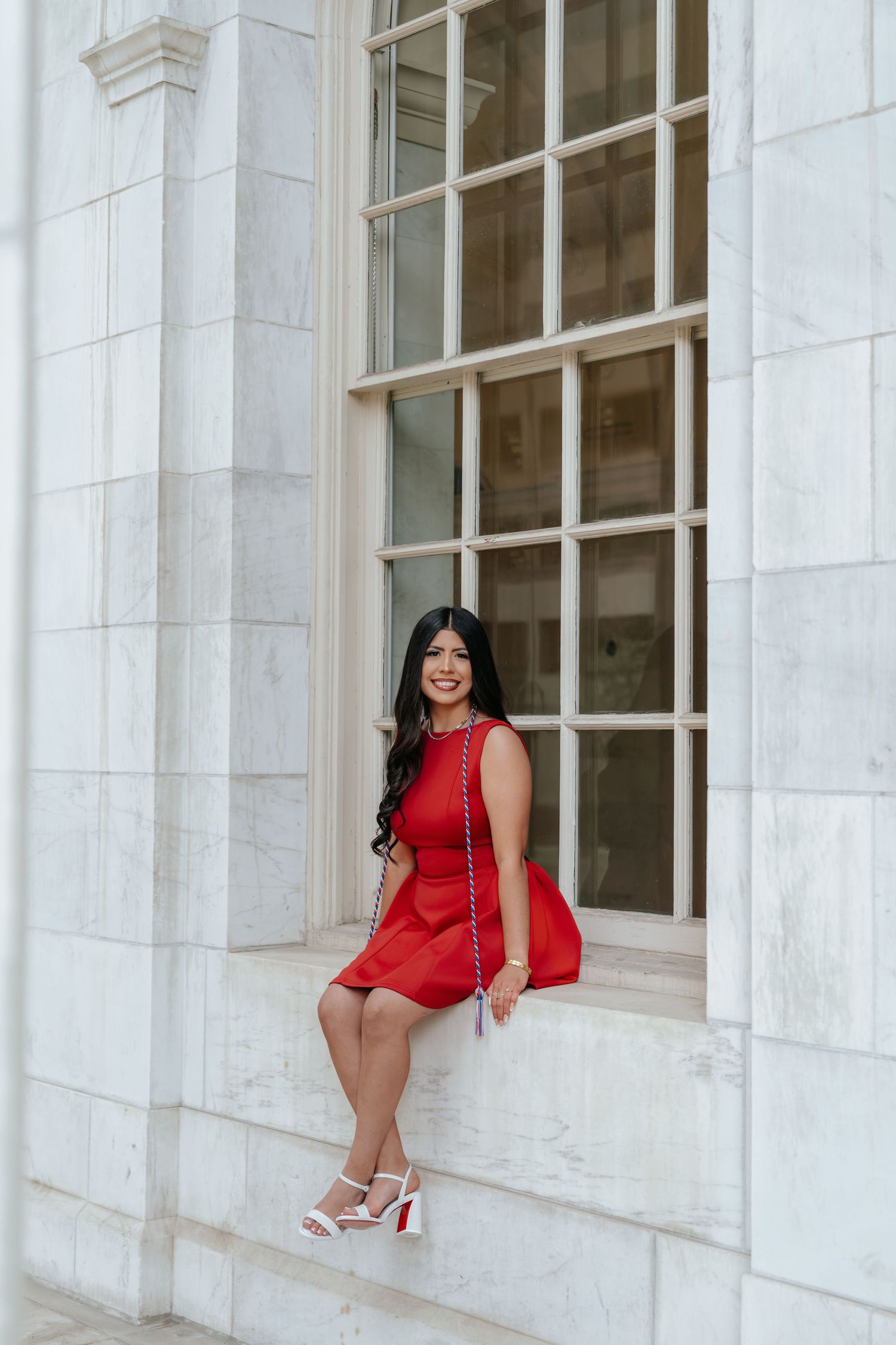 Woman in red dress sitting on marble windowsill of classical stone building, smiling at camera.