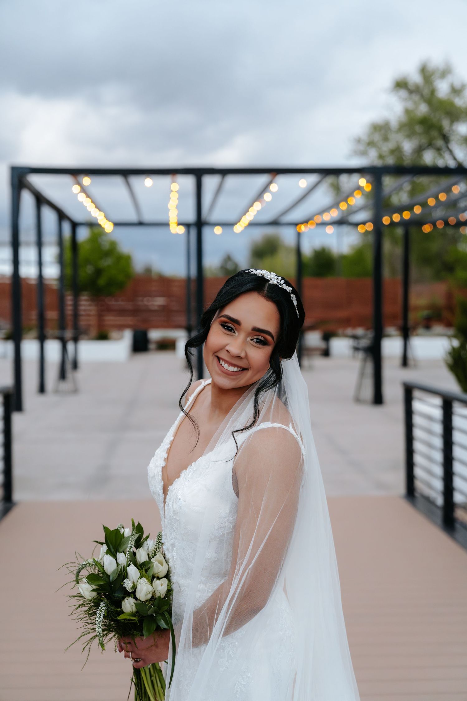 Beautiful bride in lace wedding dress and veil holds white tulip bouquet on elegant outdoor venue deck.