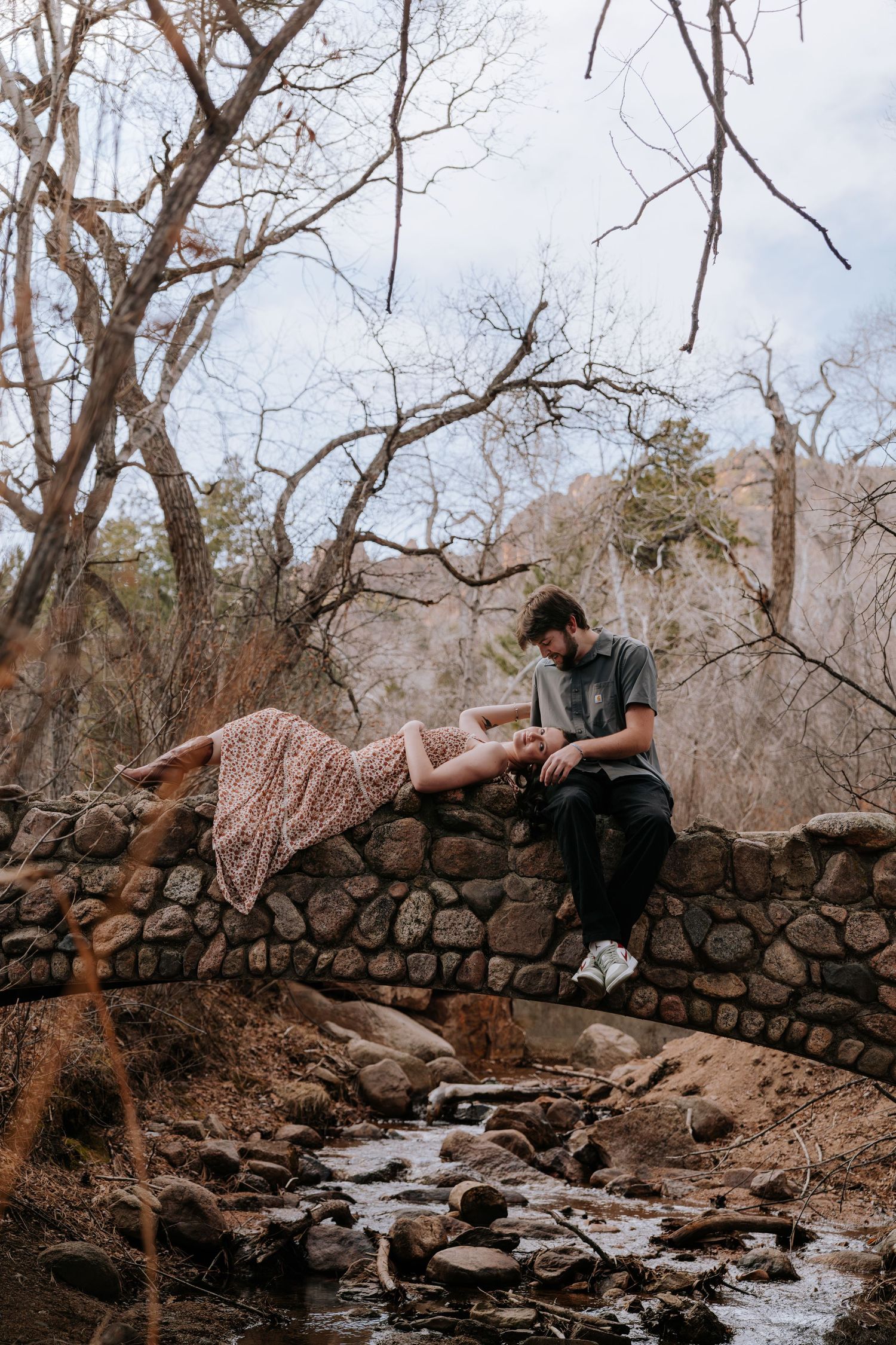 Woman in floral dress lying on stone bridge wall beside seated man near rocky creek.