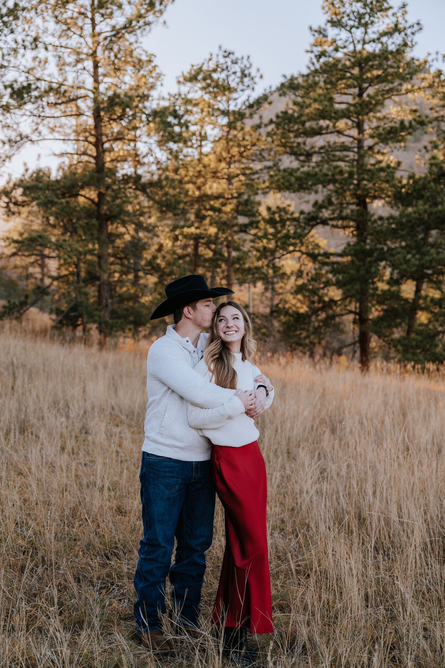 Man in cowboy hat embracing woman in red pants in a golden field with pine trees at sunset.