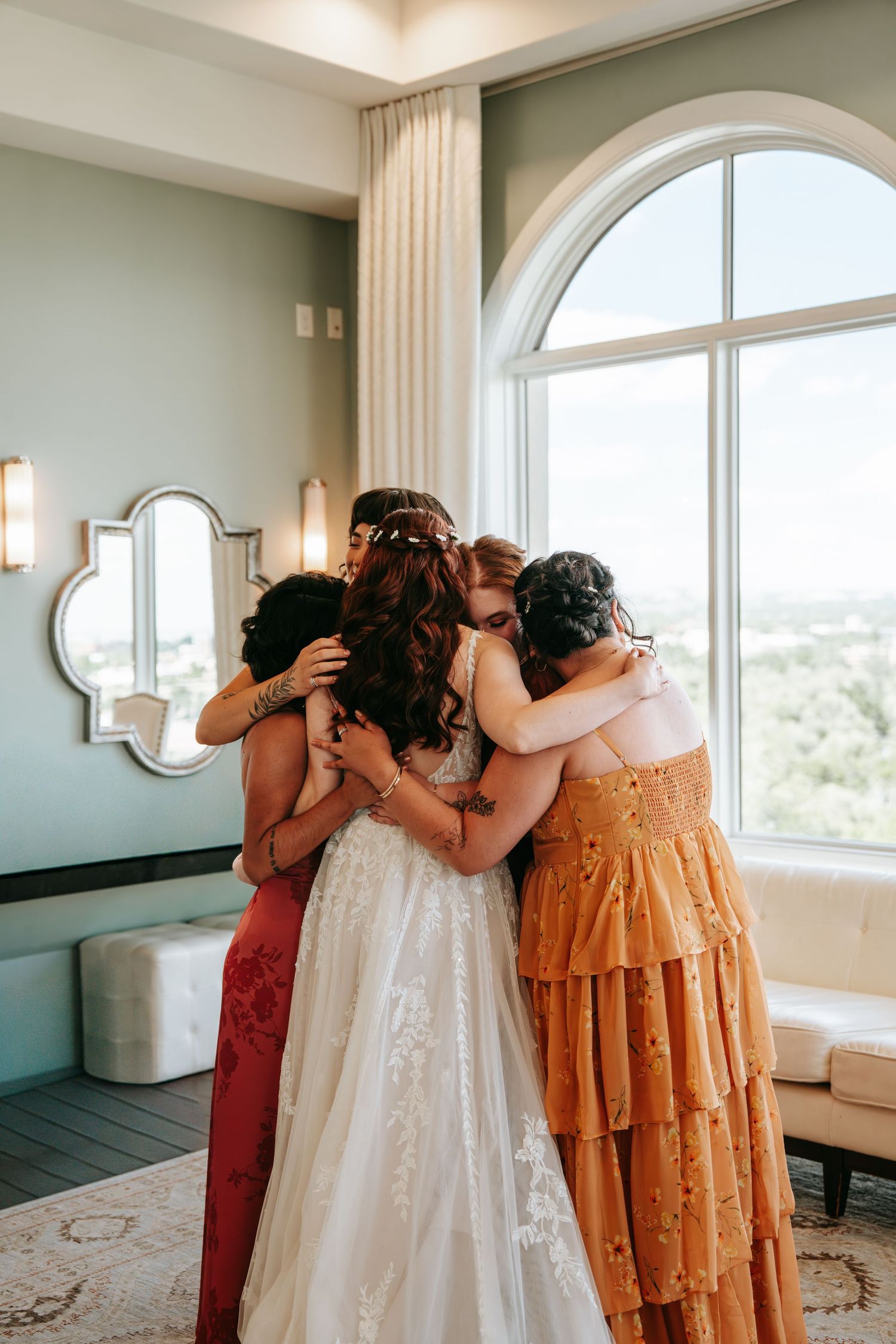 Bride in lace gown shares a heartfelt group hug with bridesmaids near a large arched window in bridal suite.