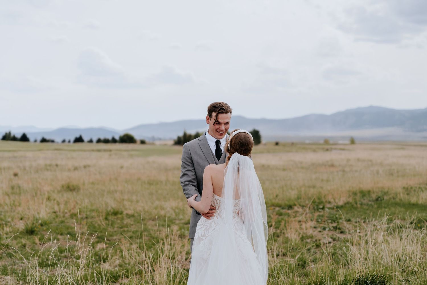 Groom in gray suit gazes lovingly at bride in white veil and lace dress in an open mountain field.