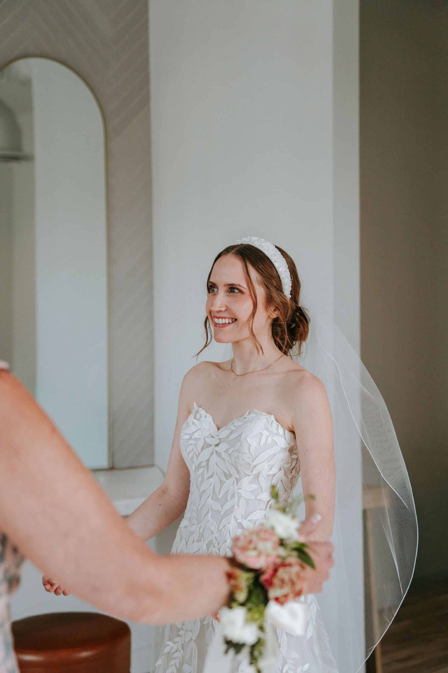 Smiling bride in strapless lace gown and veil holding bouquet in bright wedding venue.