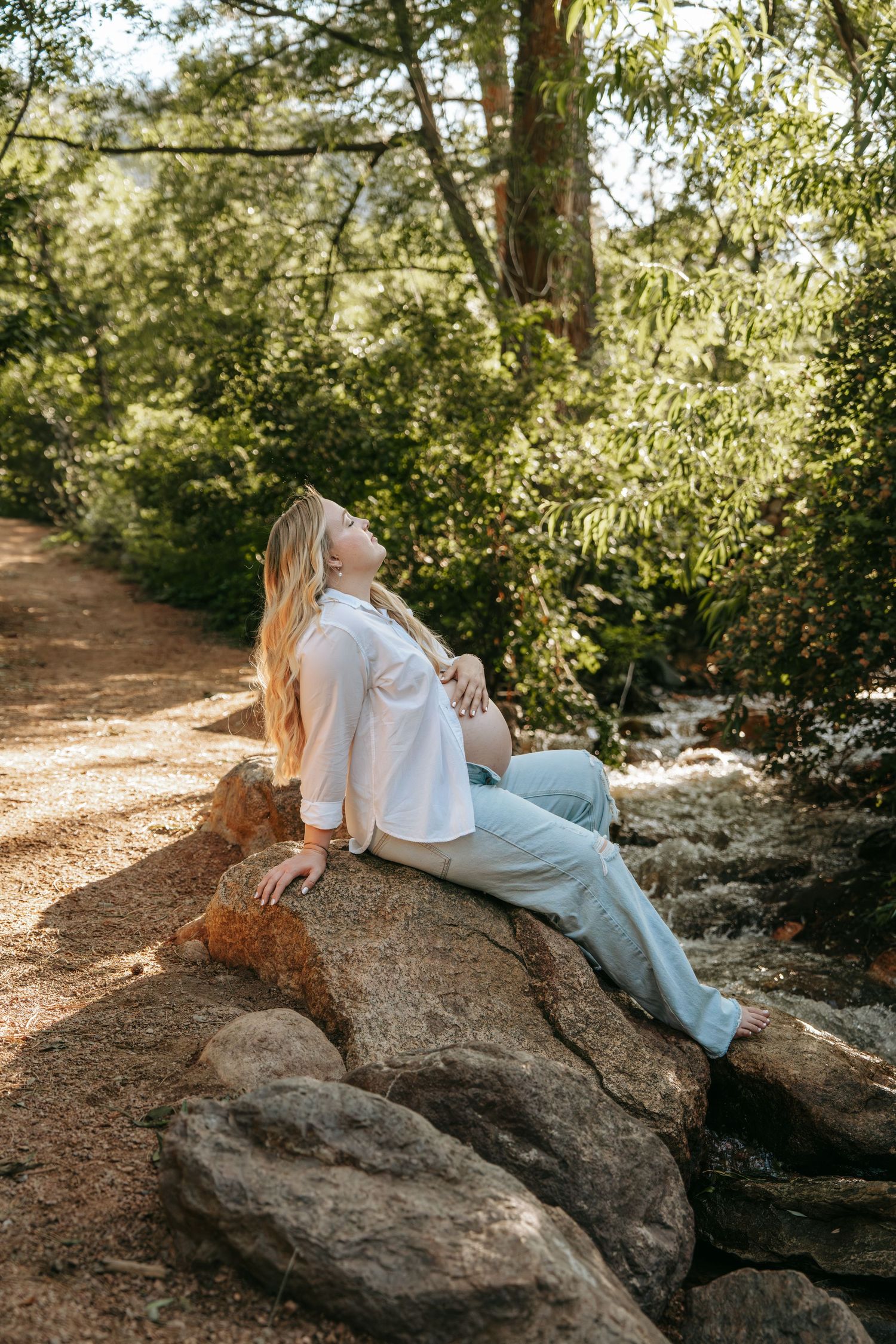 Pregnant woman reclines on rocks in a sunlit woodland, head tilted back peacefully.