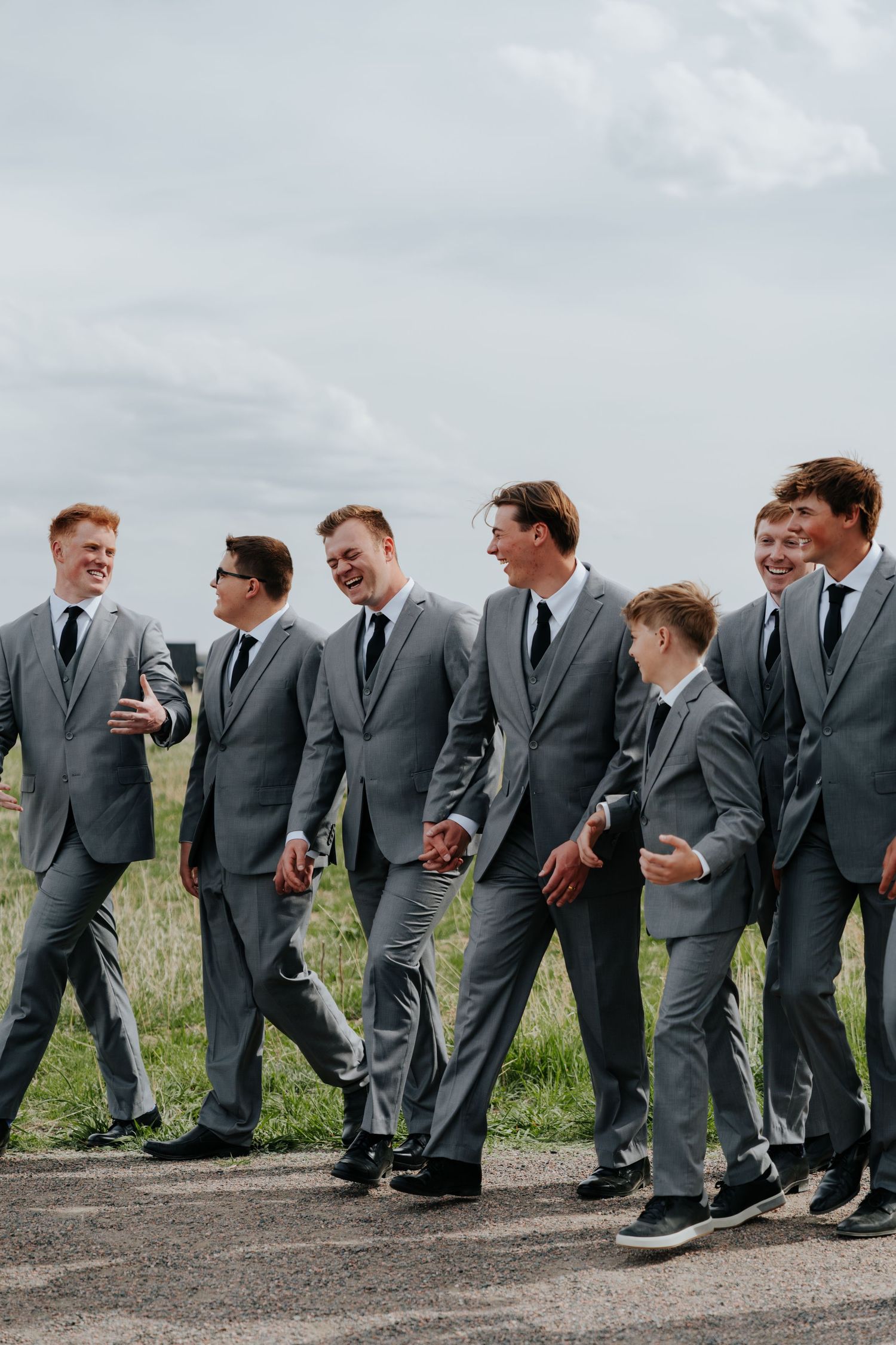 Groomsmen in matching gray suits and black ties laugh together while walking outdoors on a cloudy day.