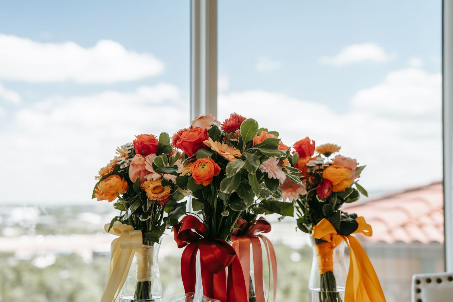 Three vibrant orange and red floral bouquets tied with colorful ribbons displayed on a windowsill with blue sky.