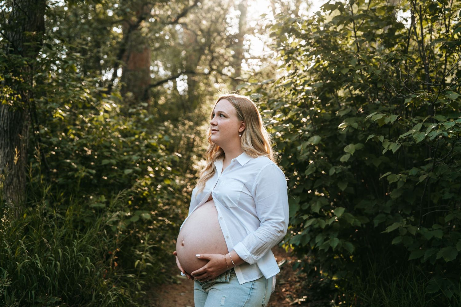Smiling pregnant woman cradling her bare baby bump on a golden sunlit forest trail.