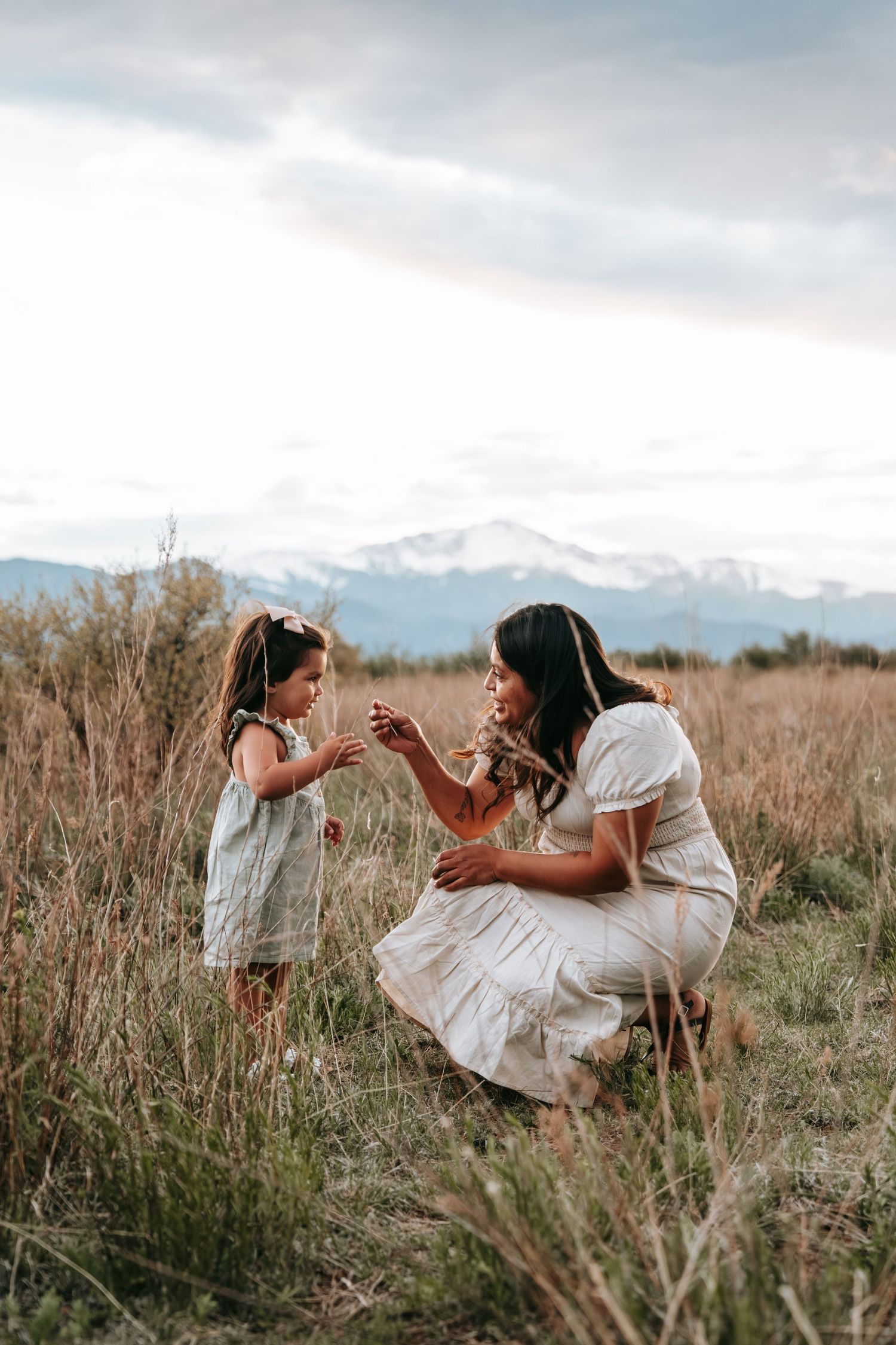 Mother in white dress crouches to interact with young daughter in grassy mountain field.