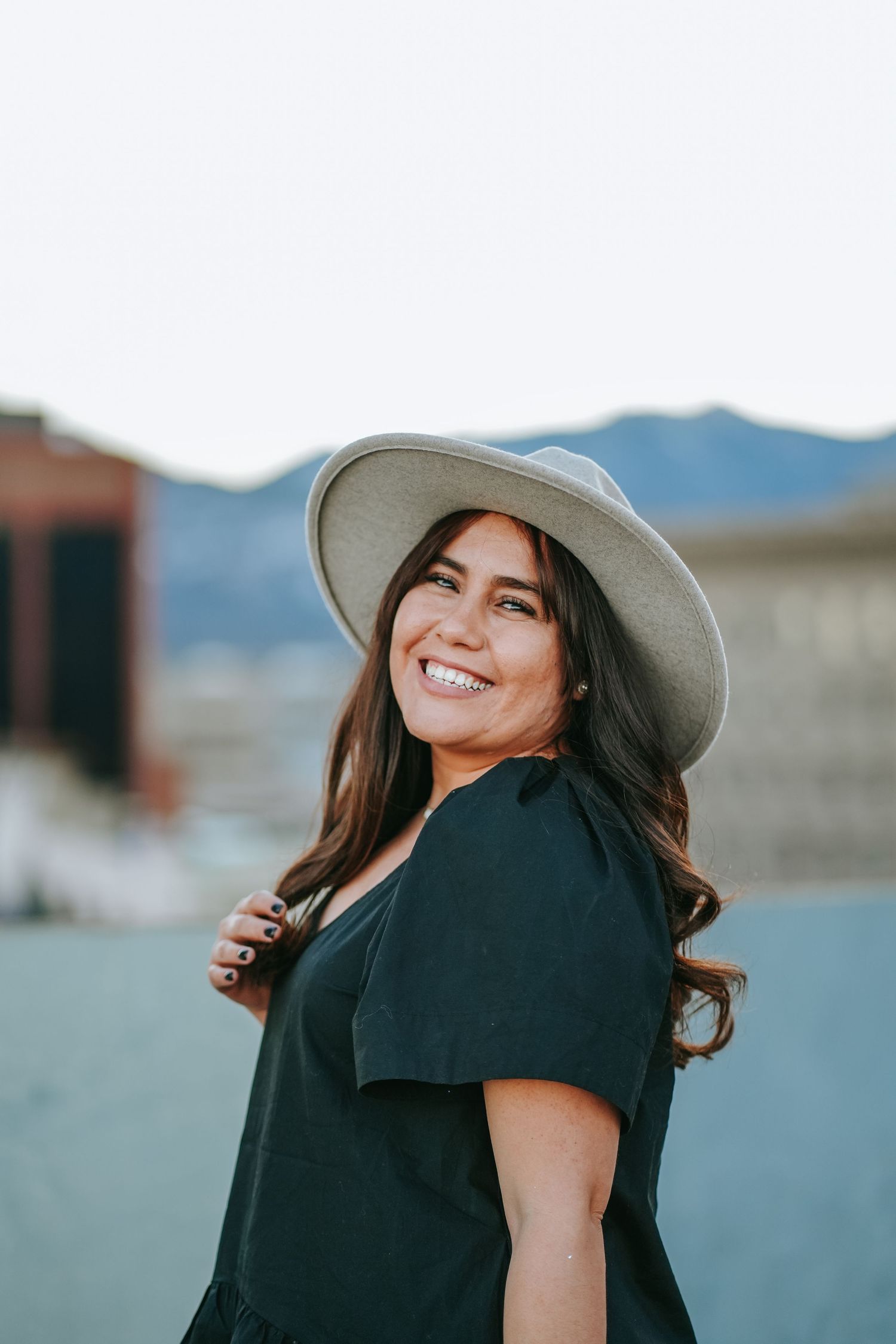 Smiling woman in a black dress and gray hat posing on a rooftop with mountains behind.