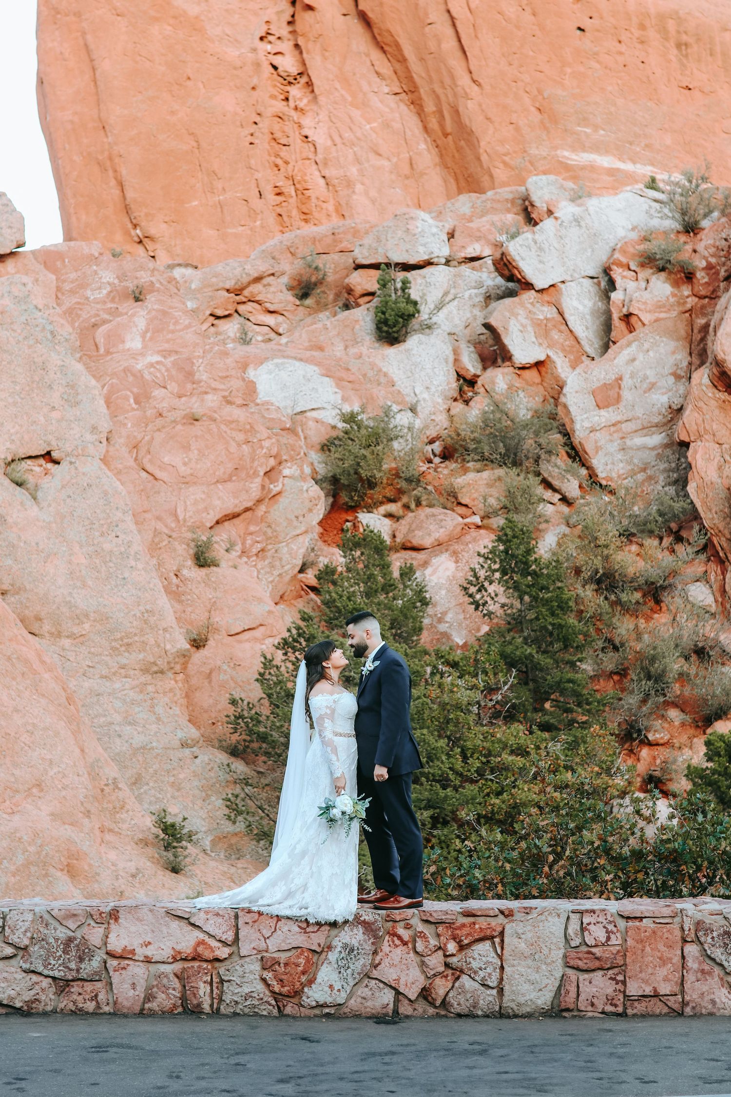 Bride and groom standing together on stone wall with dramatic red rock formations behind.