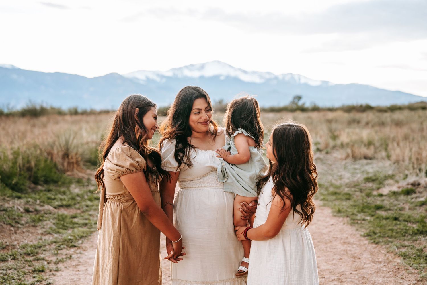 Mother in white dress holding toddler surrounded by three daughters on a scenic mountain path.