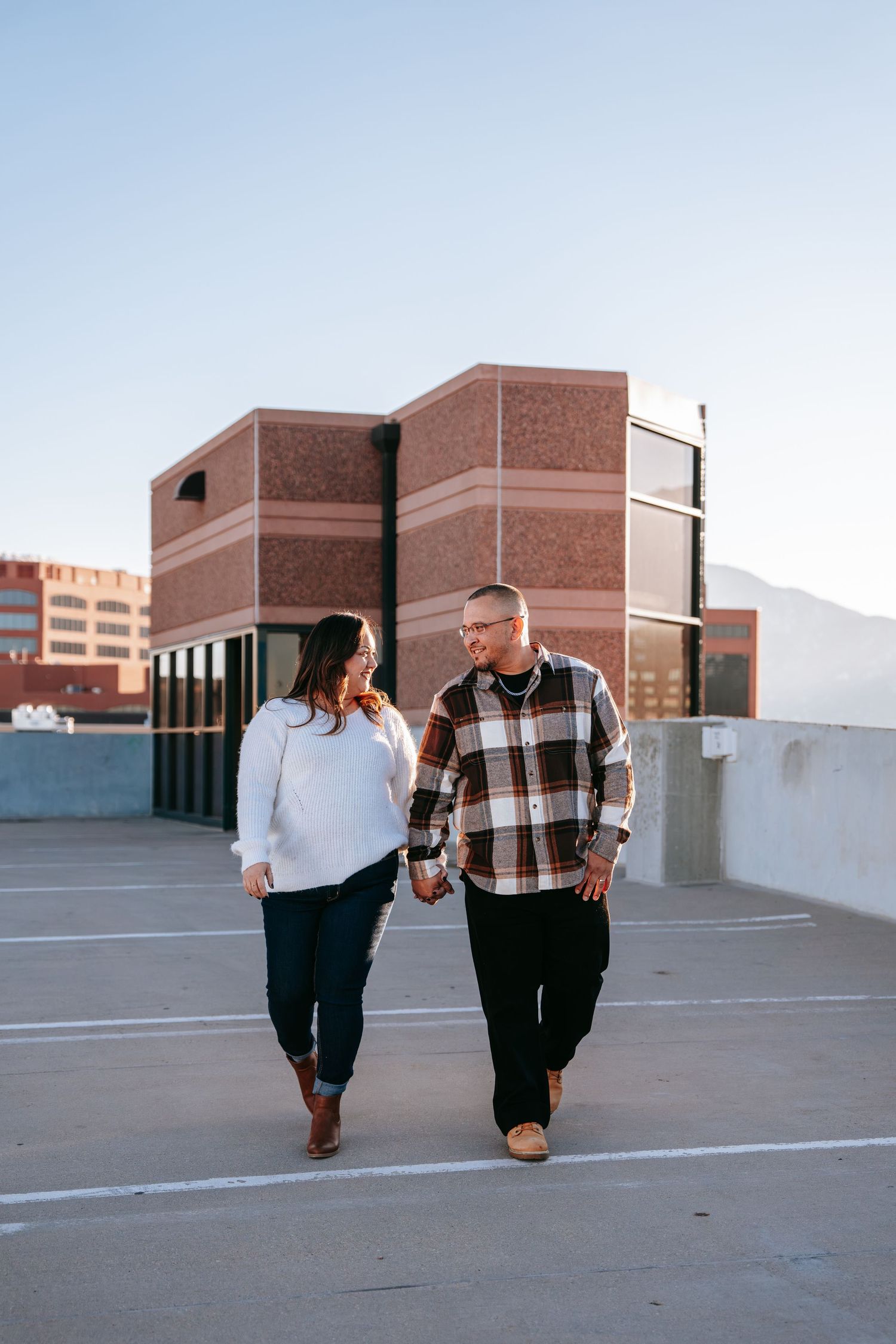 Couple holding hands and smiling while walking on rooftop parking lot at sunset.