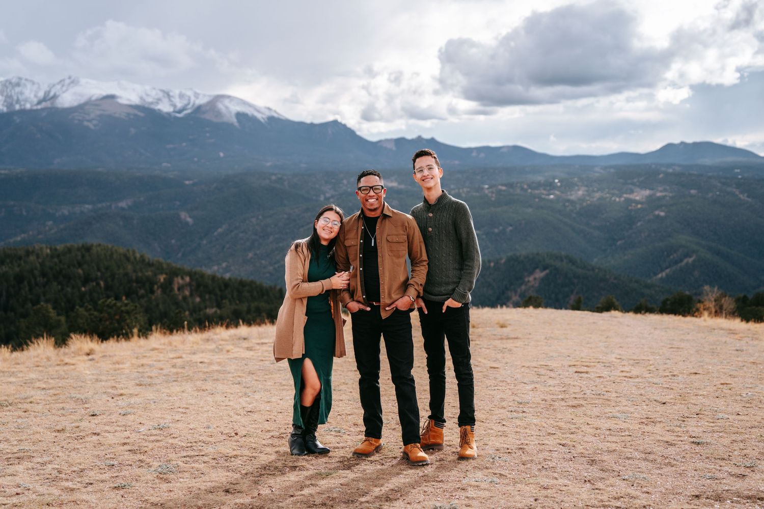Three family members smiling together on a mountain overlook with snow-capped peaks in the background.