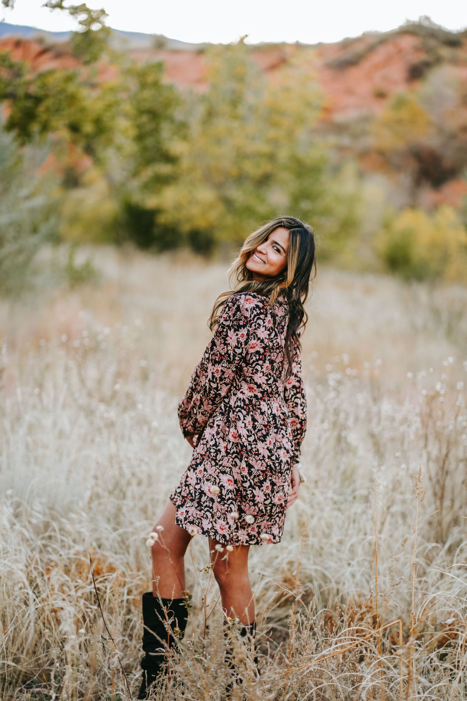 Woman in floral mini dress standing in tall dry grass field with red rocks behind.