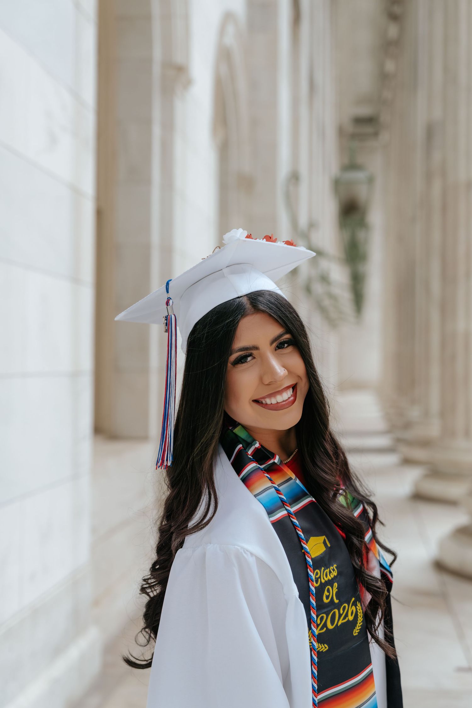 Smiling graduate in white cap and gown with colorful serape stole, Class of 2026, posing between stone columns.