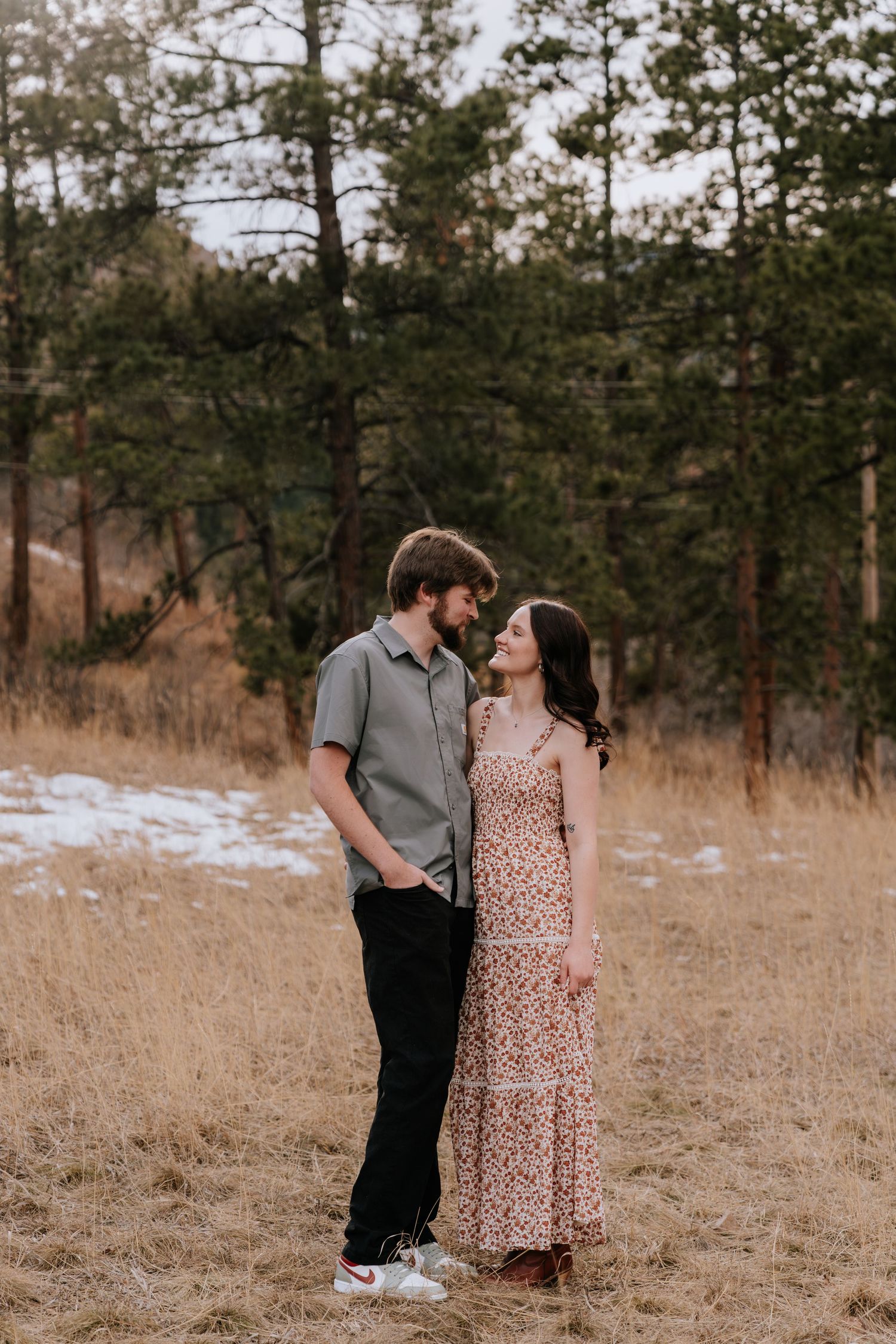 Young couple gazing at each other in golden meadow with pine trees and snow in the background.