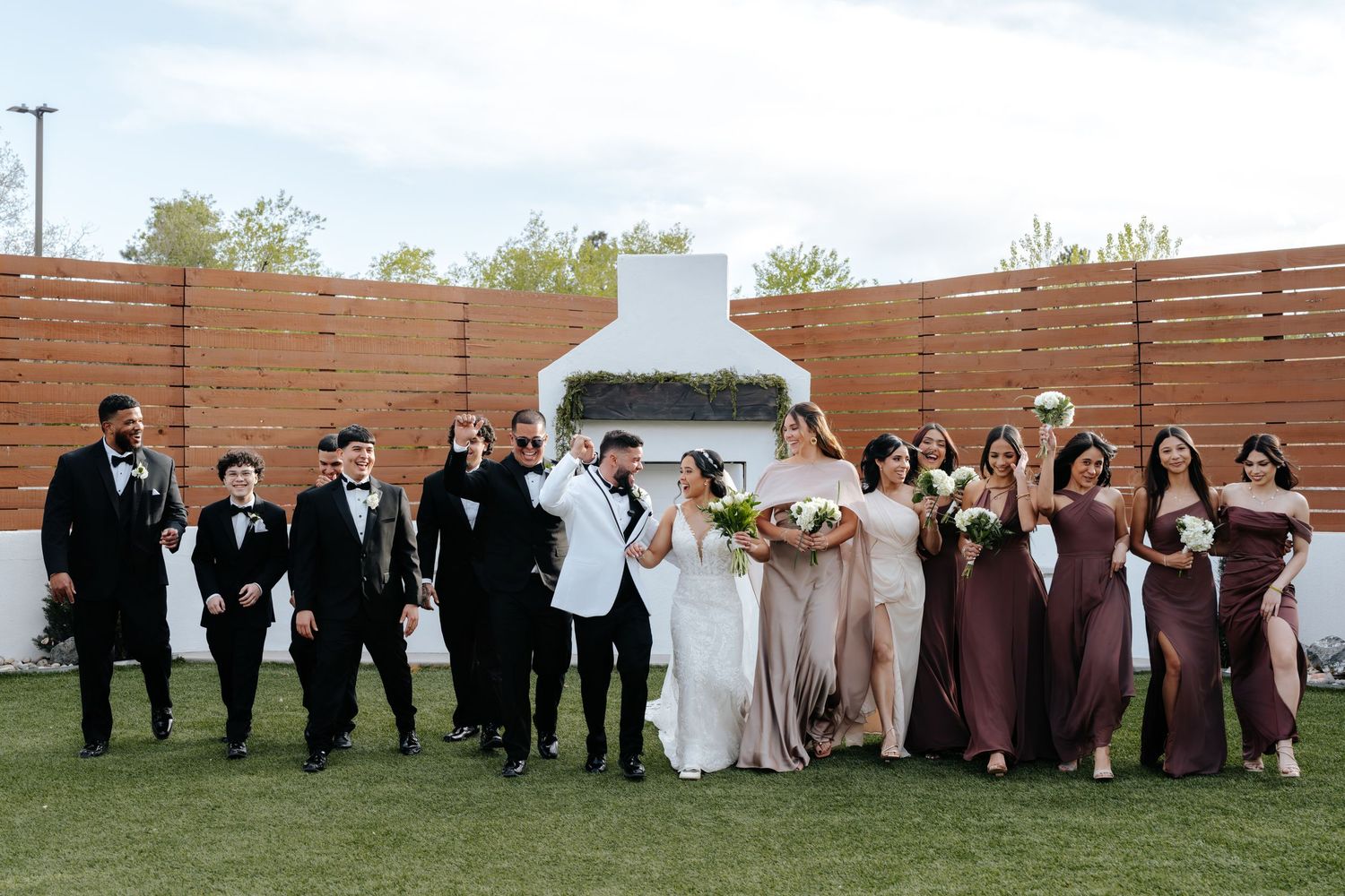 Wedding party celebrating outdoors with groomsmen in tuxedos and bridesmaids in mauve gowns near white fireplace.