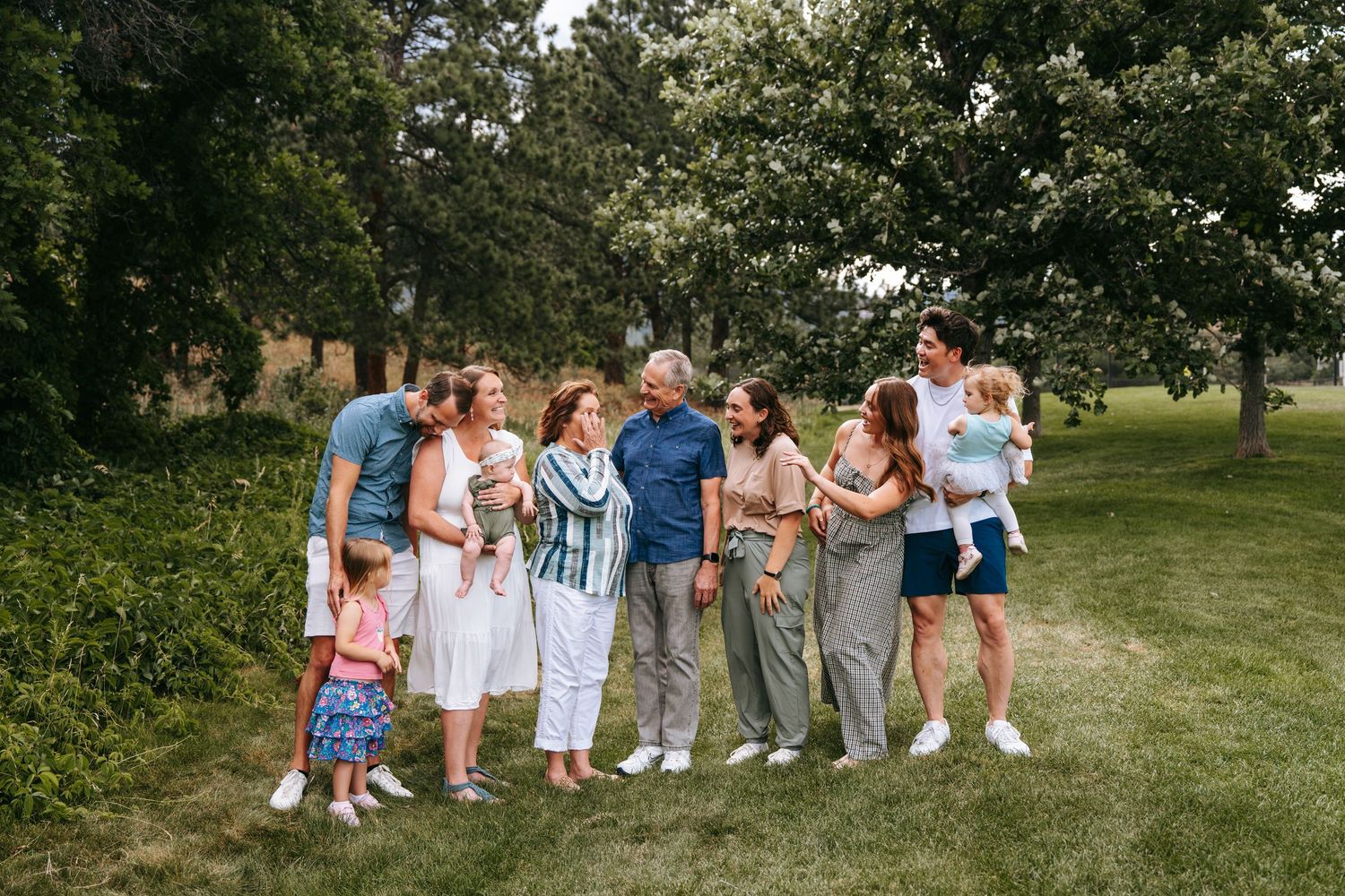 Multi-generational family gathering outdoors on green lawn surrounded by lush trees on a sunny day.