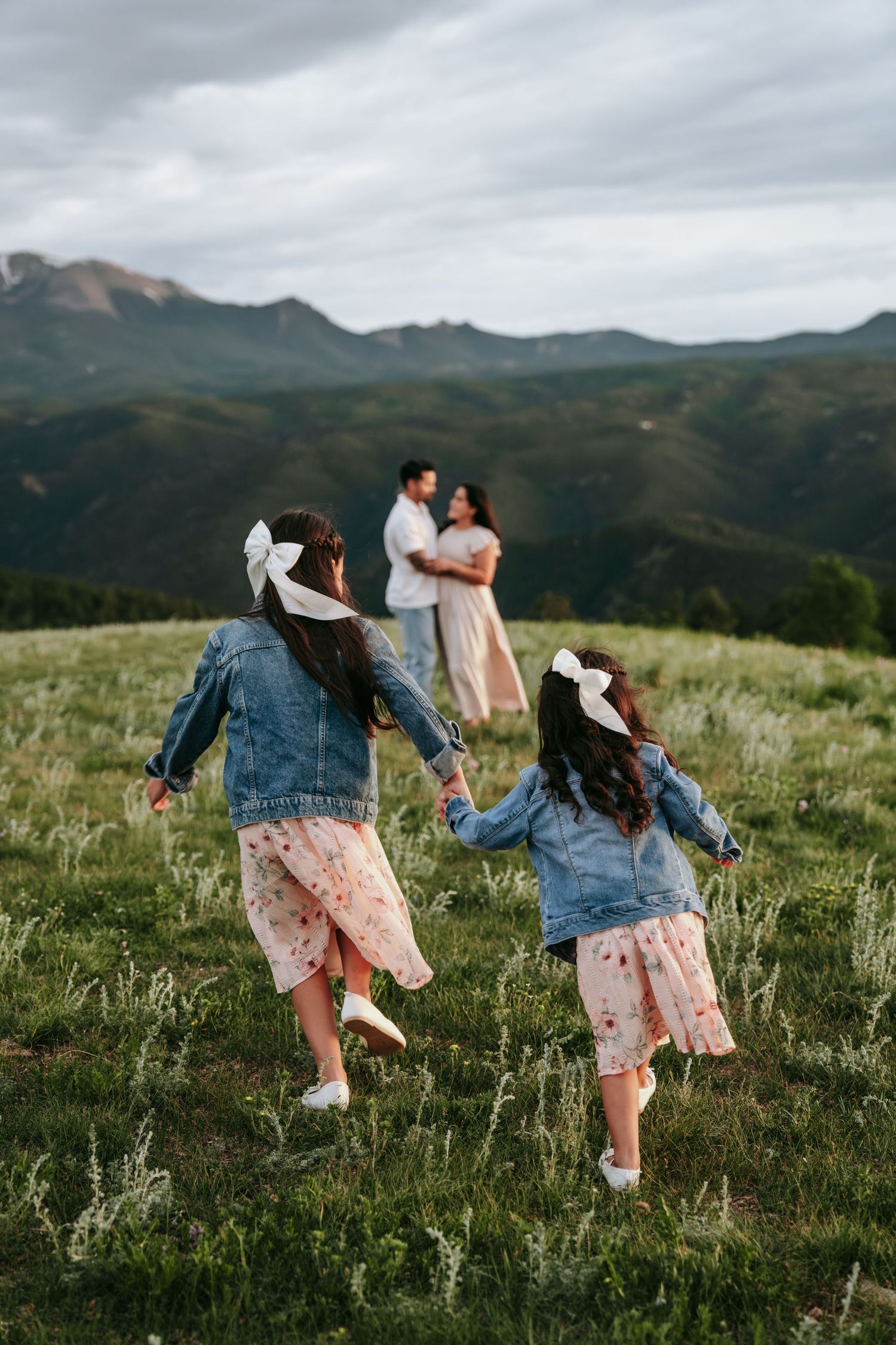 Two girls in denim jackets and bows run toward their embracing parents on mountain meadow.