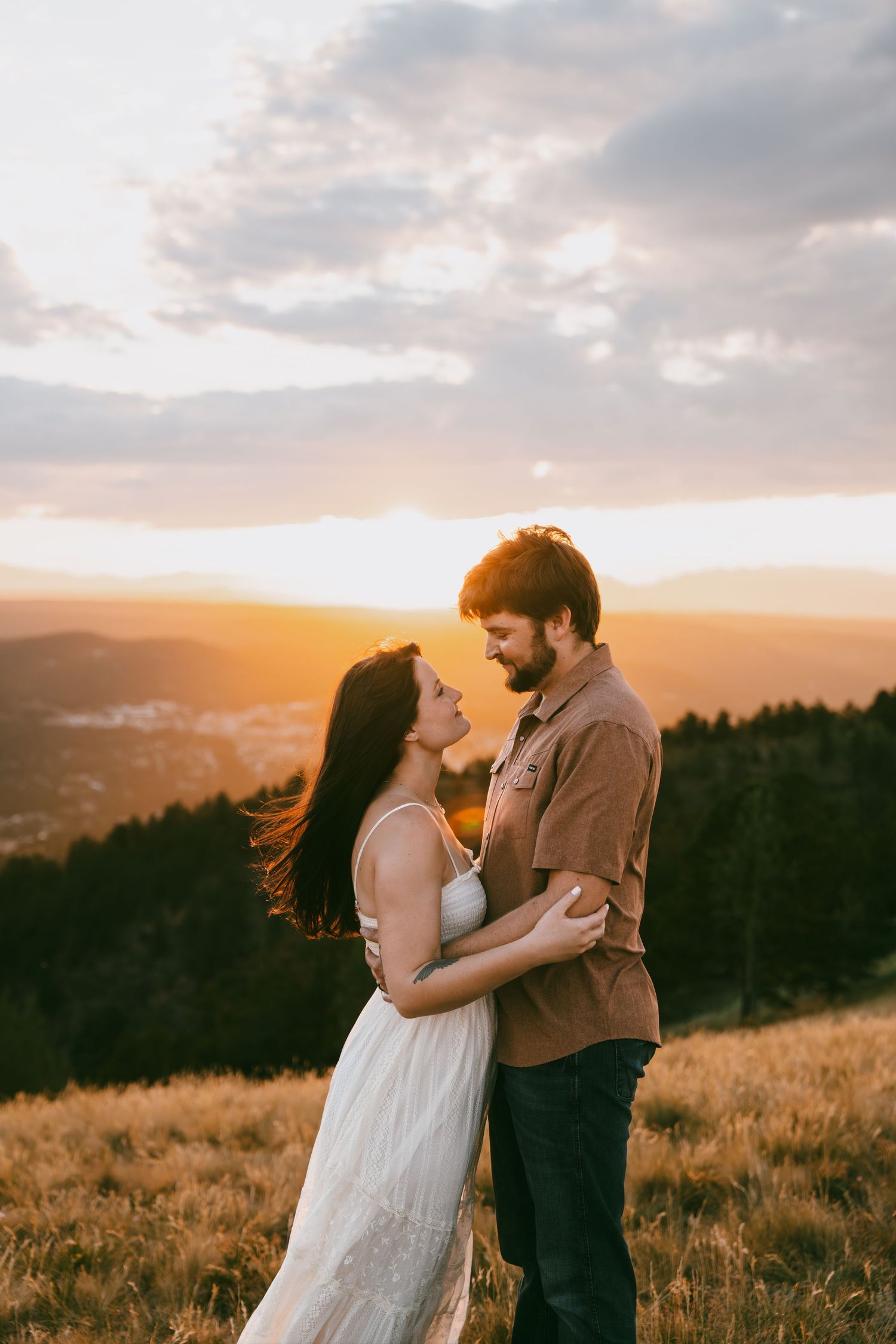 Couple embracing at golden sunset on a mountain meadow, woman in white dress and man in brown shirt sharing a tender moment.