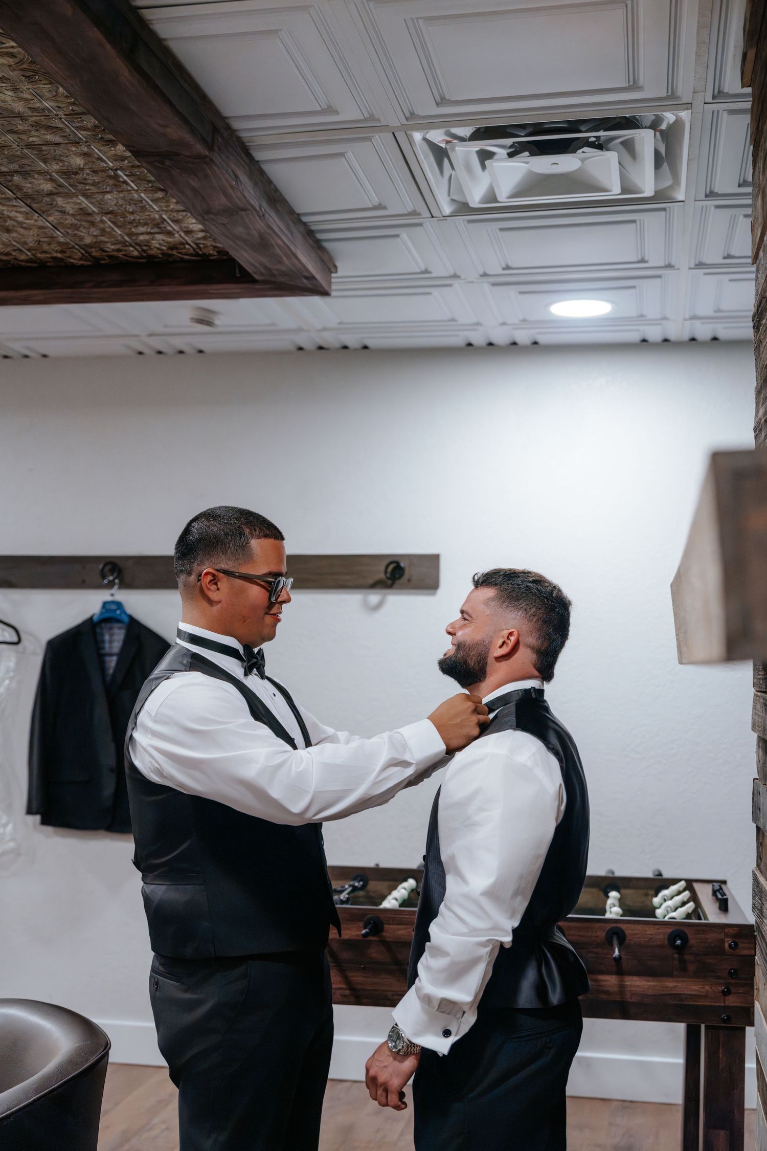 Two groomsmen in black vests and bow ties help each other get dressed in a stylish wedding preparation room.
