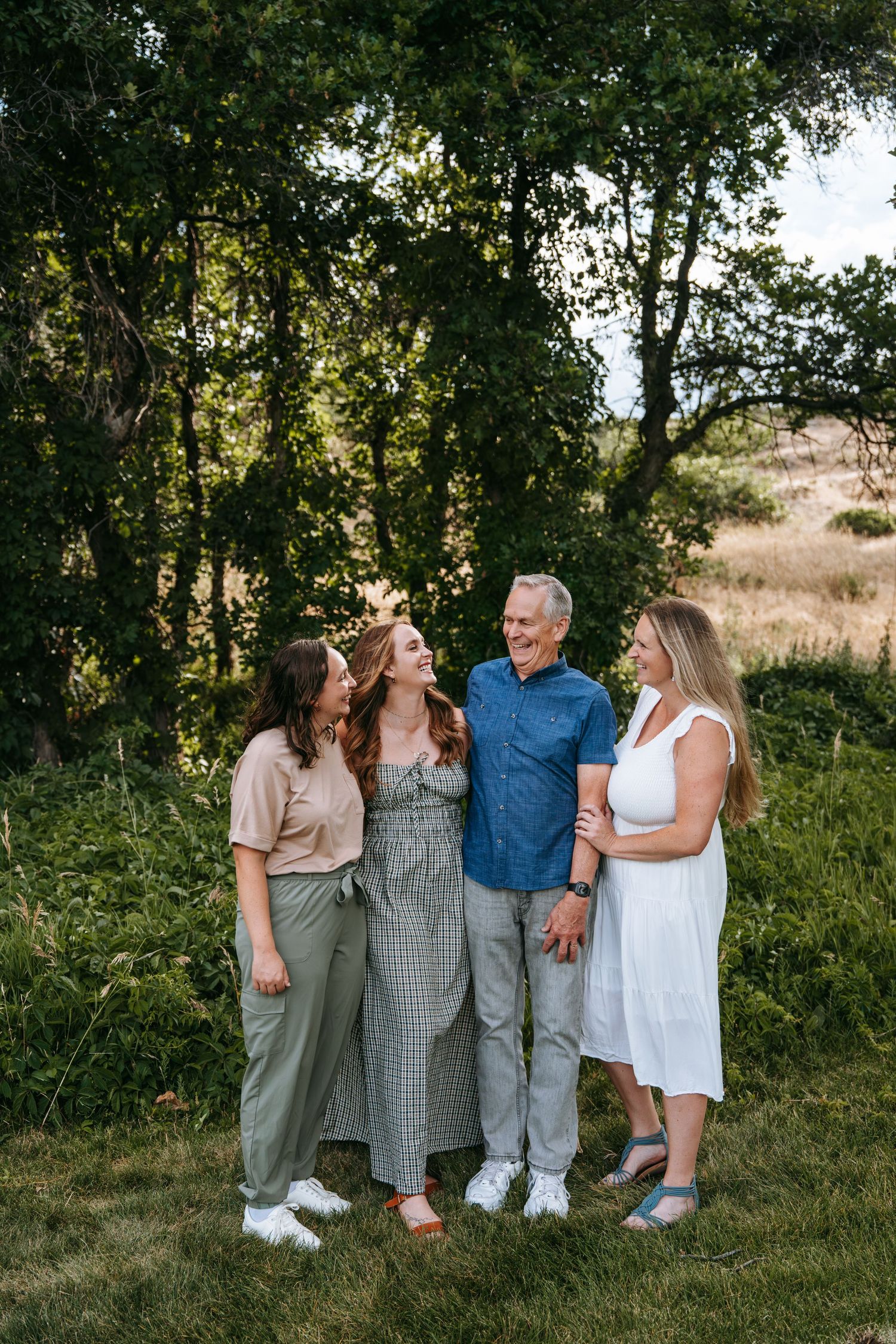 Family of four smiling and laughing together outdoors in a lush green park setting during a portrait session.