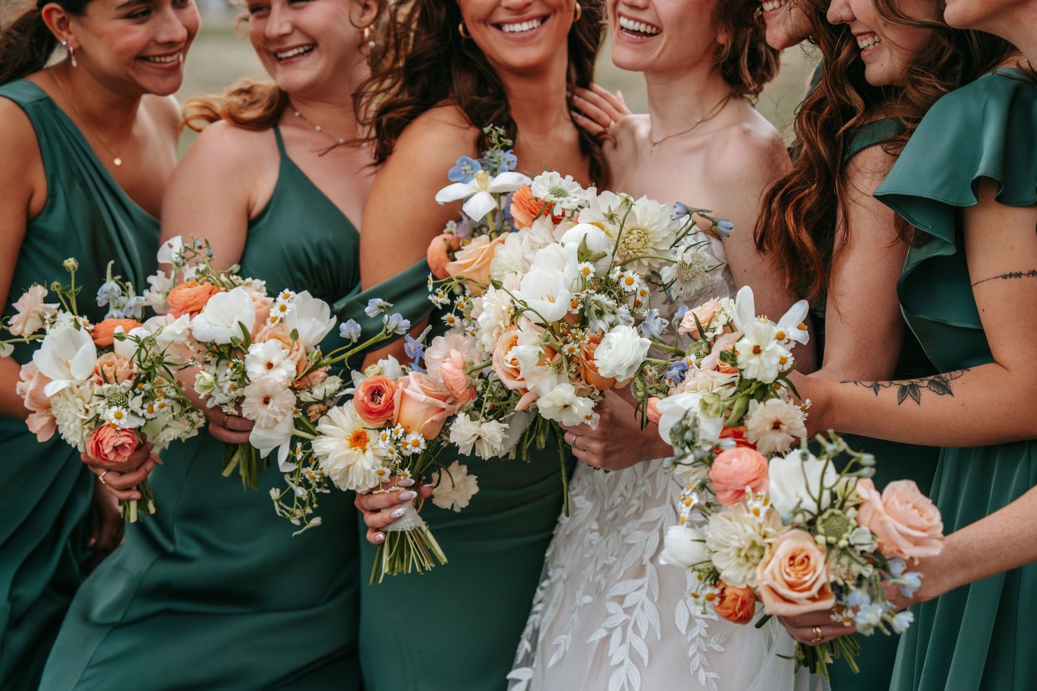 Bride and bridesmaids in emerald green dresses holding peach and white wedding bouquets while smiling.