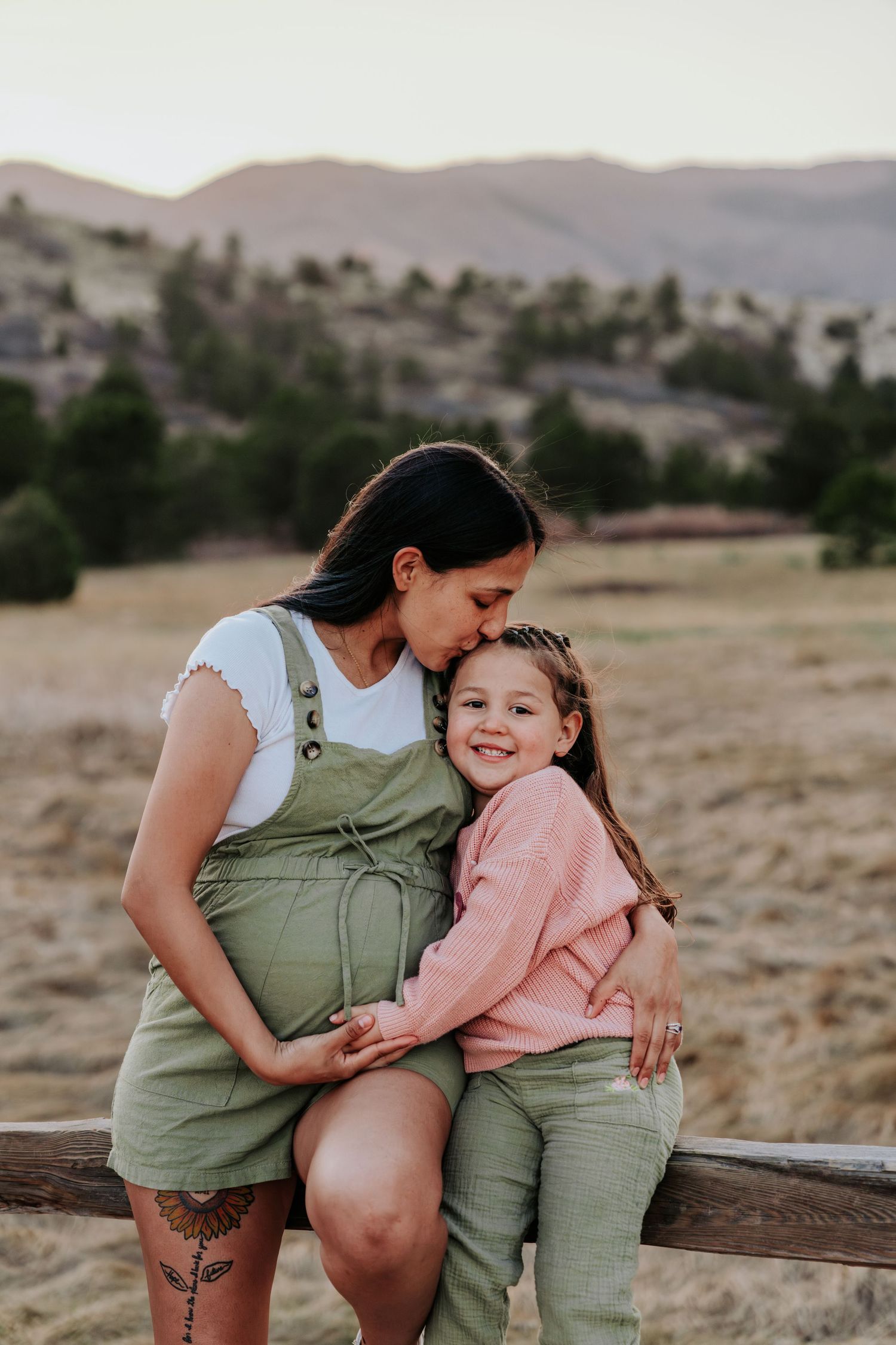Pregnant woman in green overalls kissing young girl's cheek outdoors with mountain backdrop.