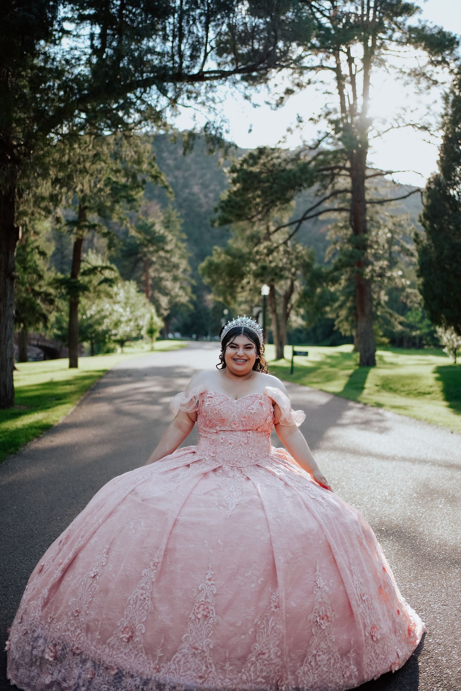 A smiling young woman in a pink quinceañera gown and tiara walks down a tree-lined path.