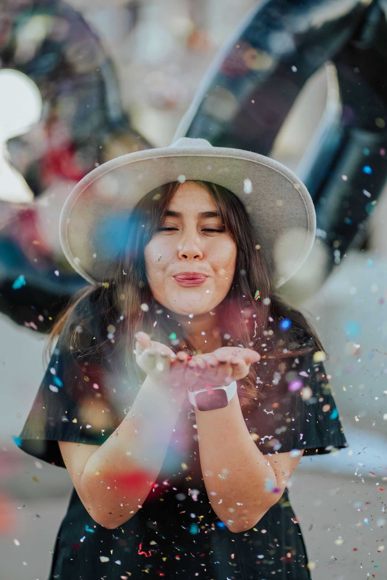 A young woman in a white hat blows colorful confetti from her hands at a birthday celebration.