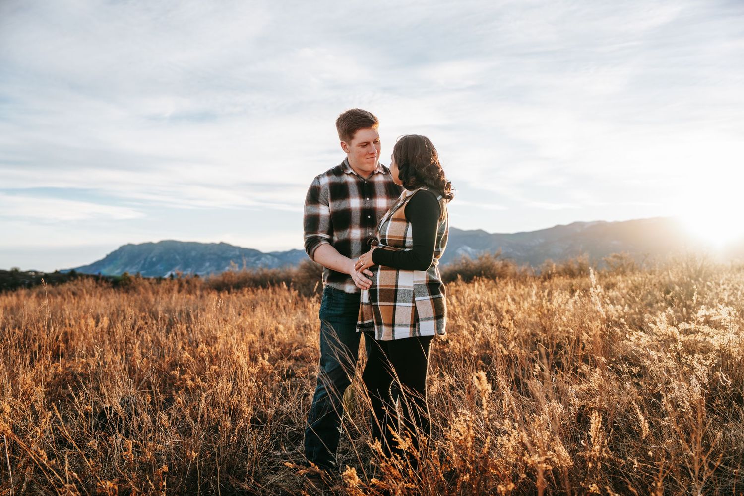 Pregnant couple embracing and sharing a kiss in a golden field with mountains at sunset.