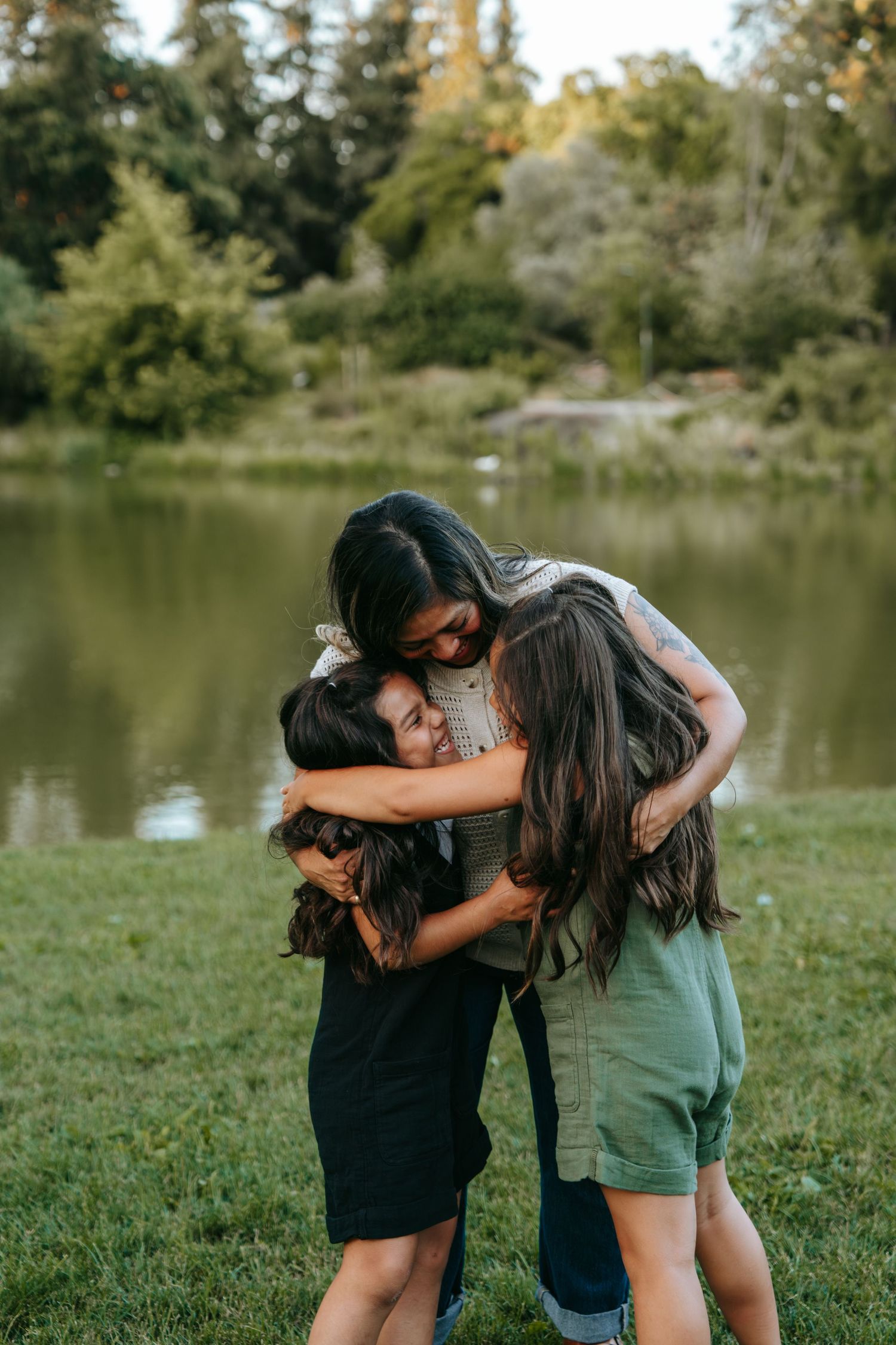 Mother sharing a warm group hug with her three children on a grassy lakeside in a park.