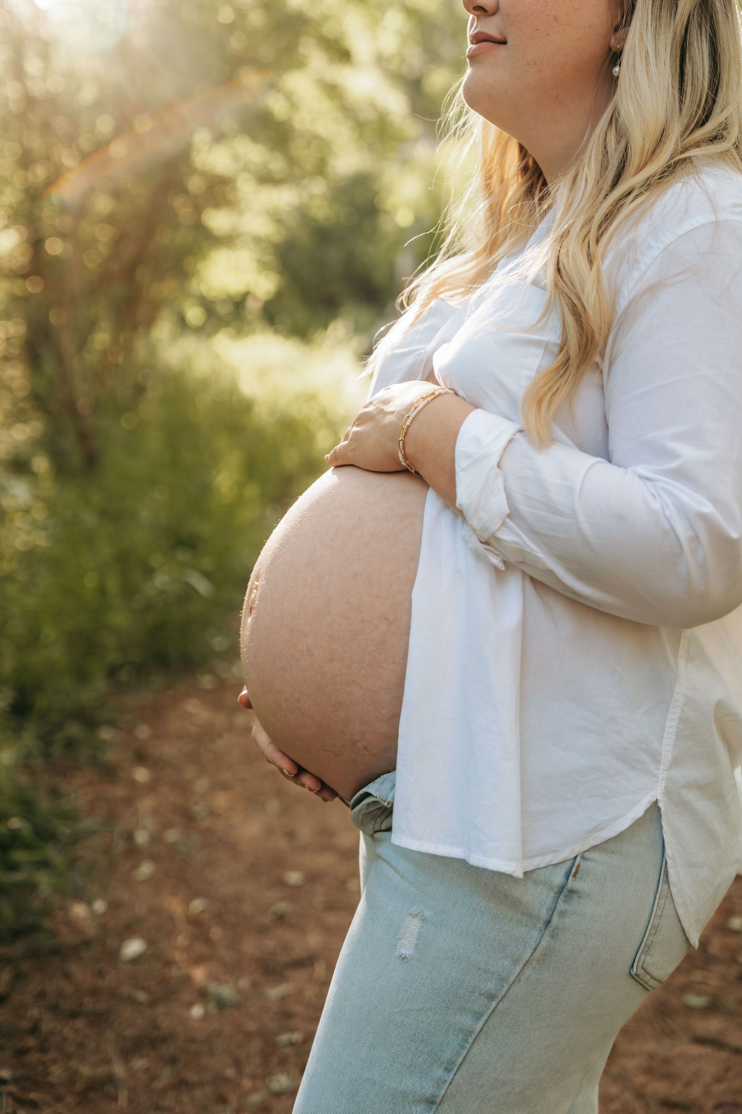 Close-up of pregnant woman in white shirt and jeans cradling her bare baby bump outdoors.