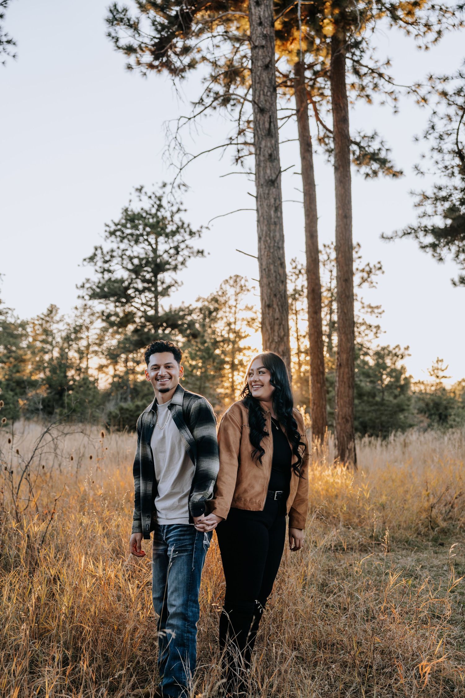 Smiling couple strolling hand in hand through a sunlit meadow surrounded by tall pine trees.