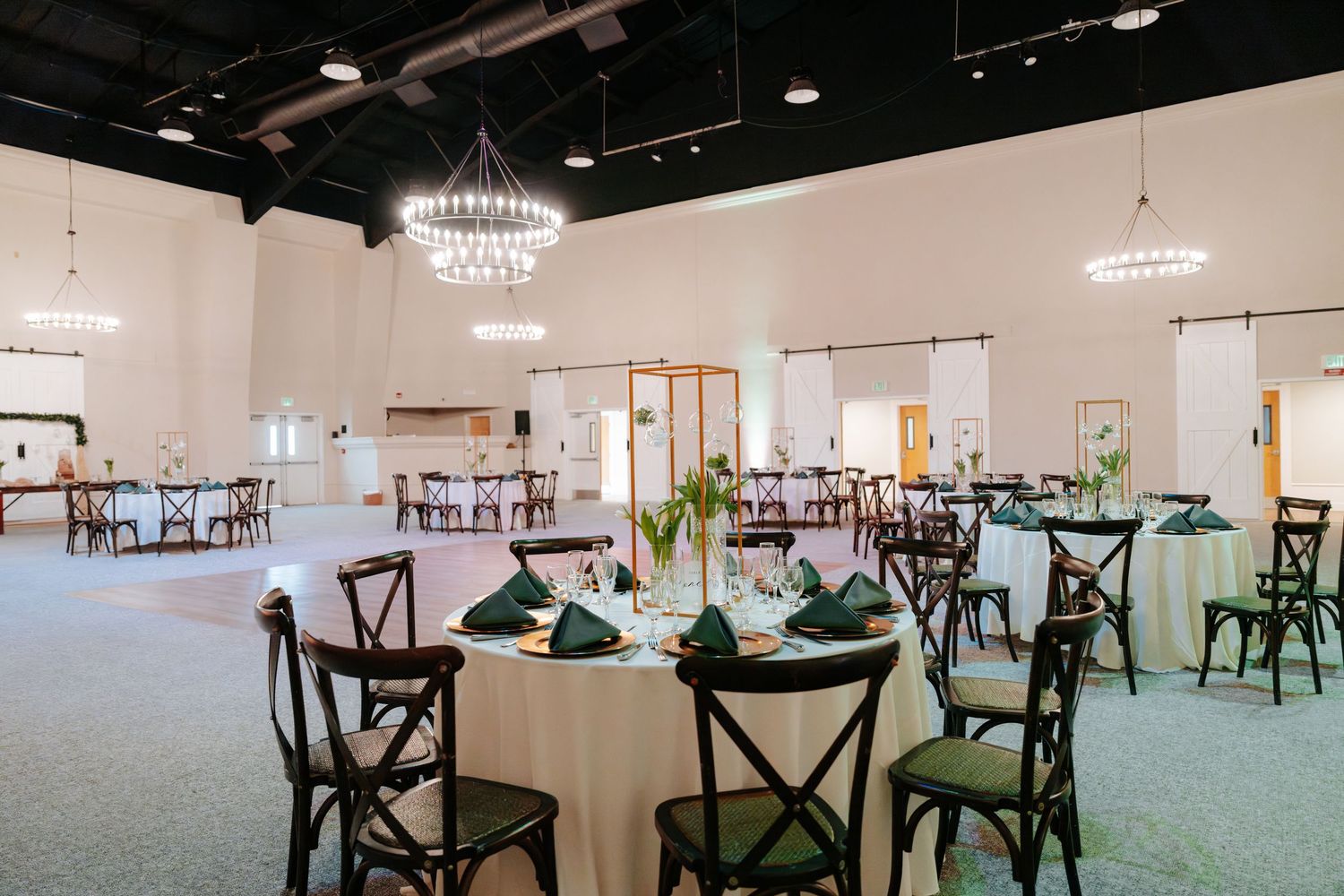 Elegant event venue with round tables, green napkins, cross-back chairs, and a crystal chandelier on white walls.