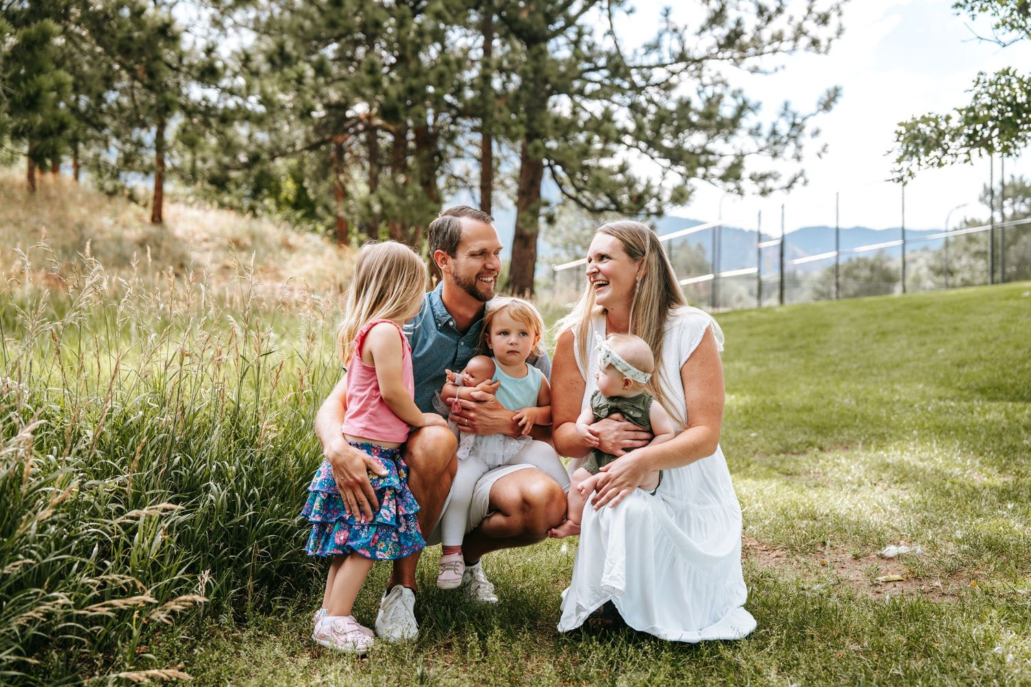 Happy family of five sitting together on green grass with pine trees and mountains in the background.