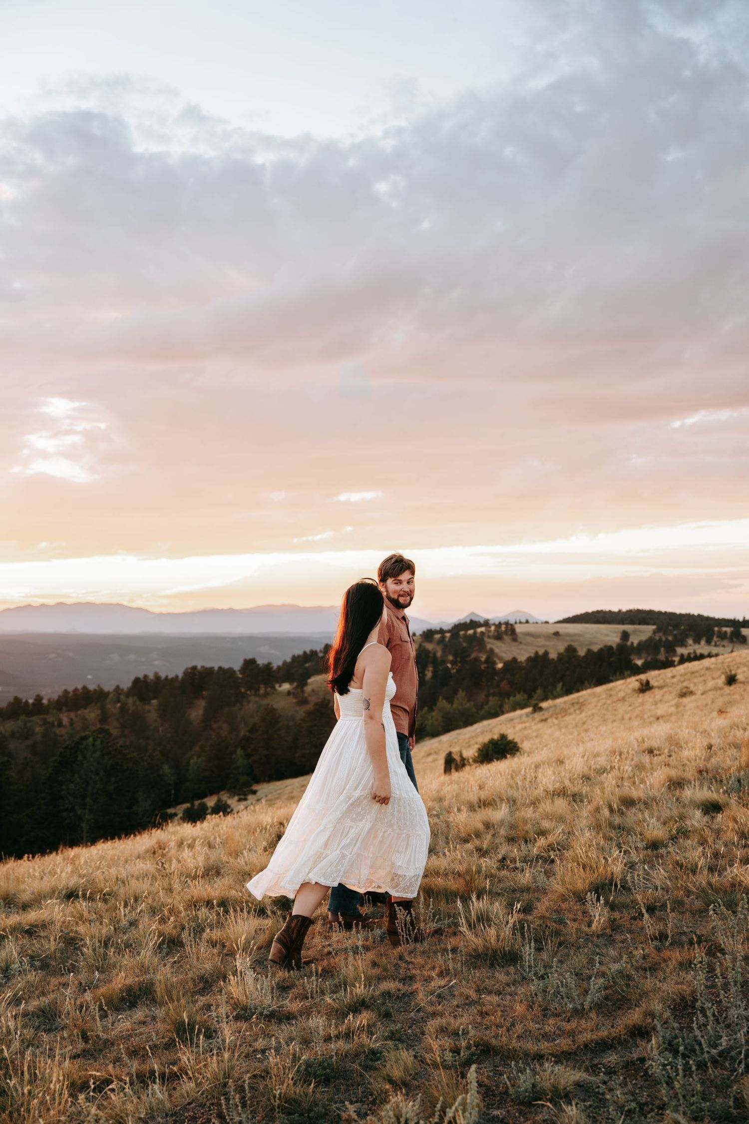 Couple in wedding attire embracing on a golden hillside at sunset with mountain views in the background.