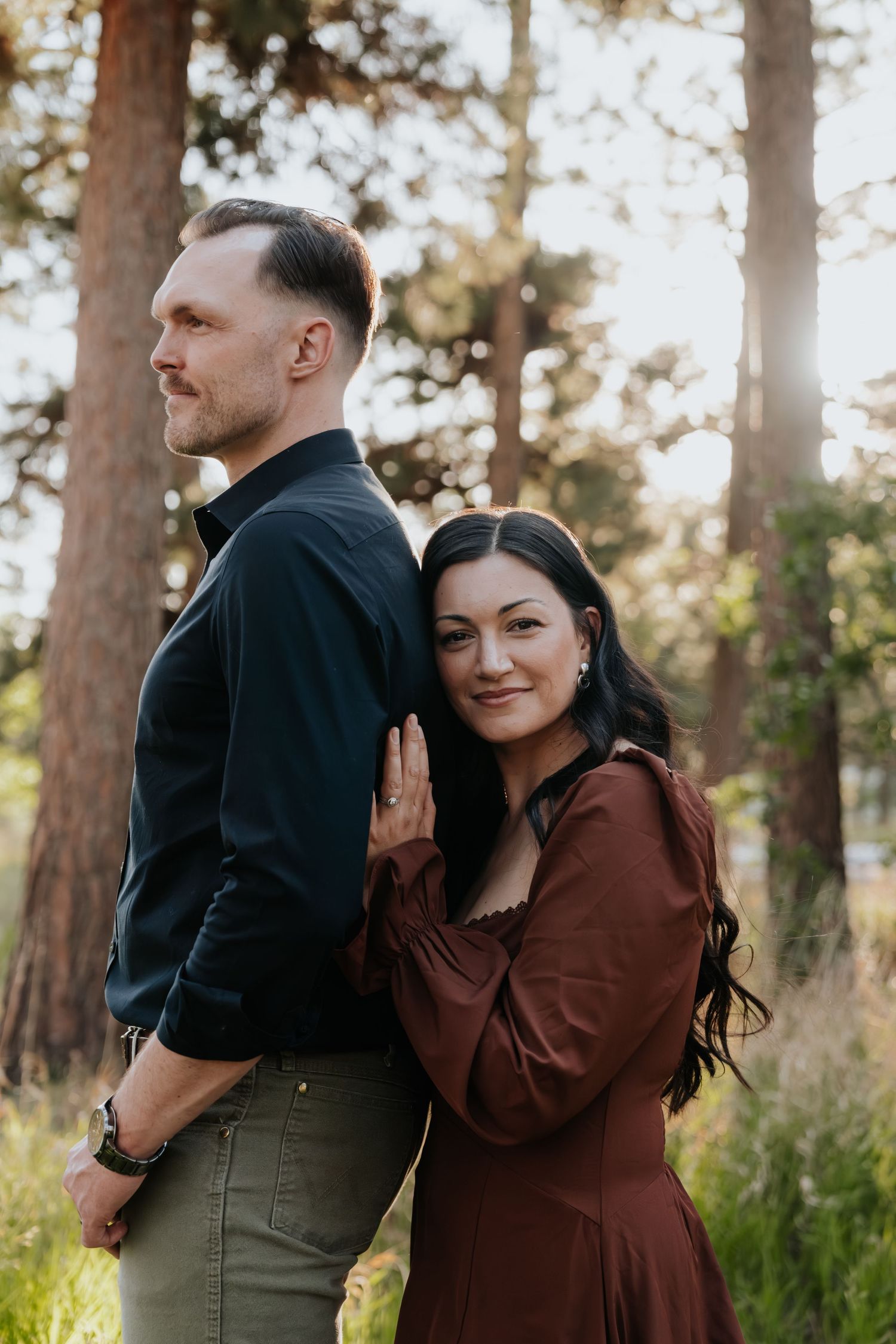 Couple poses in sunlit forest, woman in burgundy dress smiles at camera while leaning against man in dark shirt.