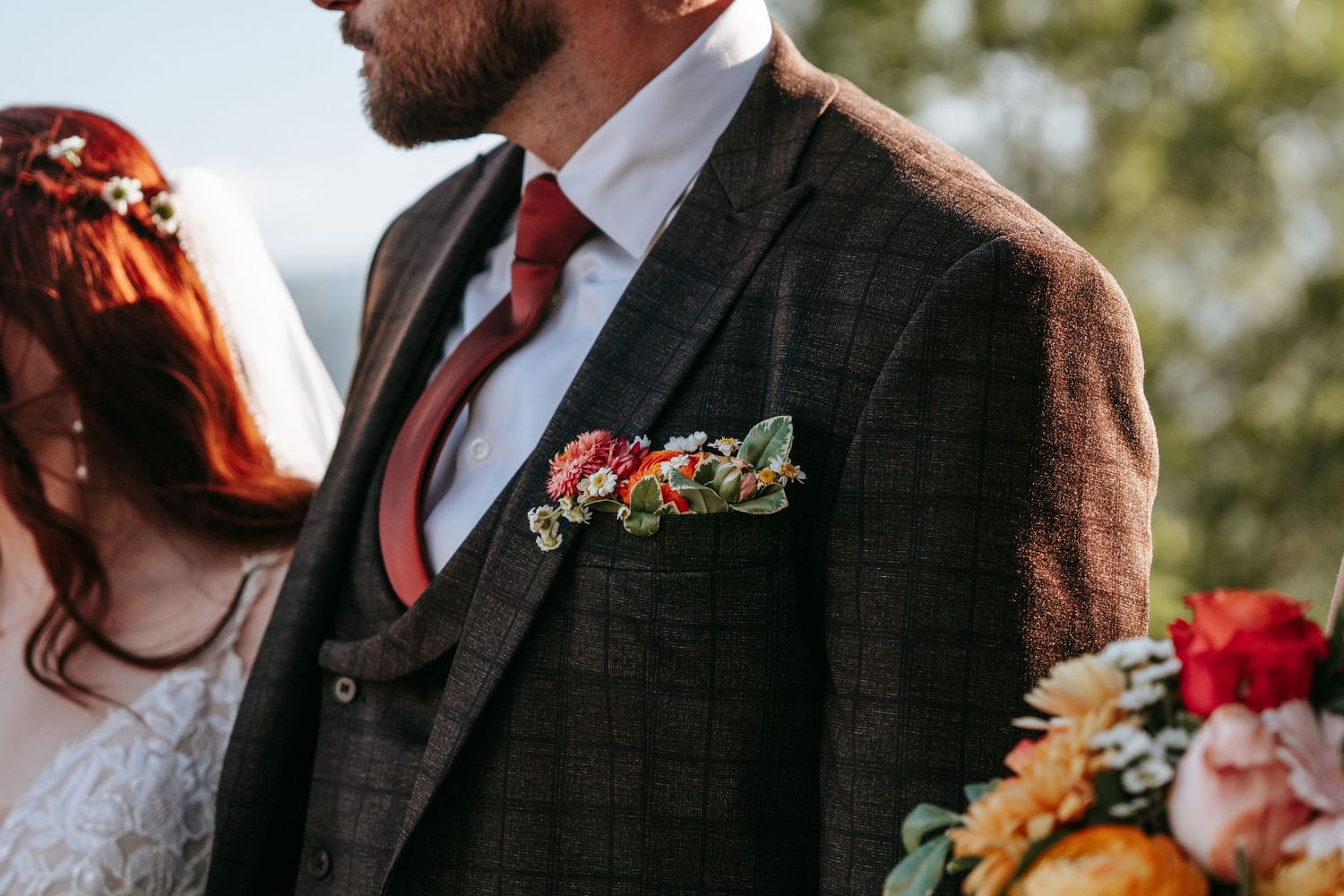 Close-up of groom in brown tweed suit with a colorful floral boutonniere and burgundy tie.