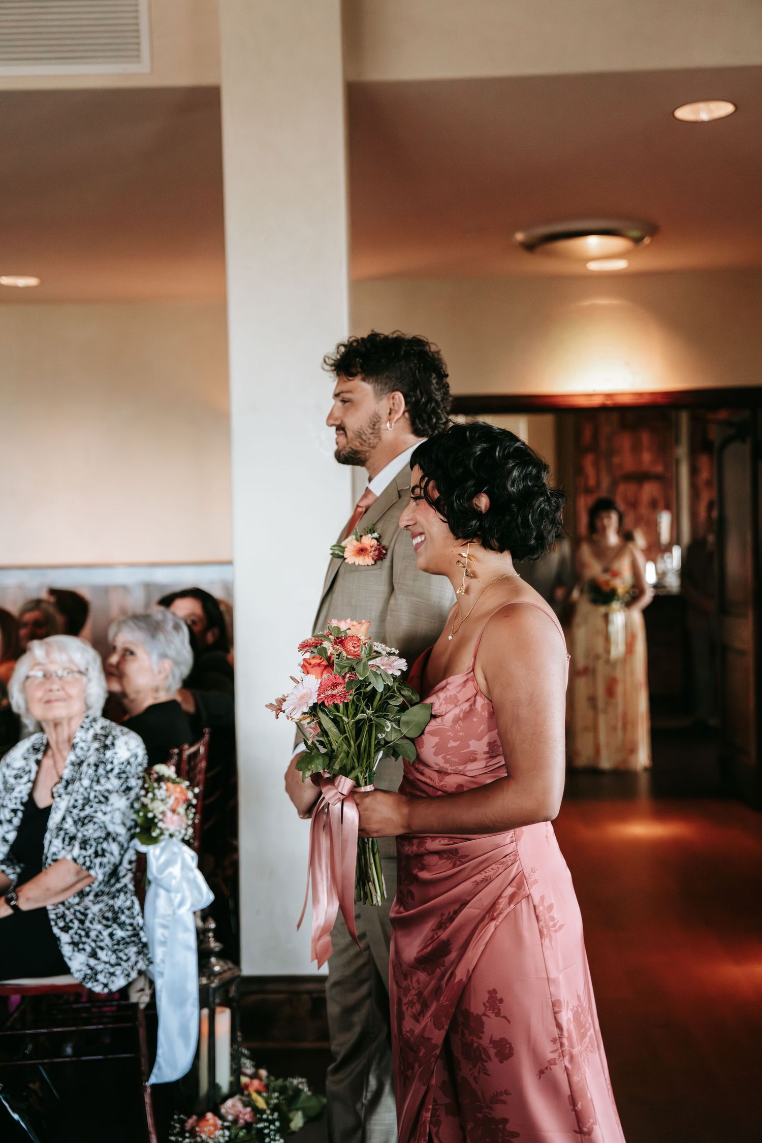 Groomsman escorts bridesmaid in pink floral dress down the aisle as wedding guests look on warmly.
