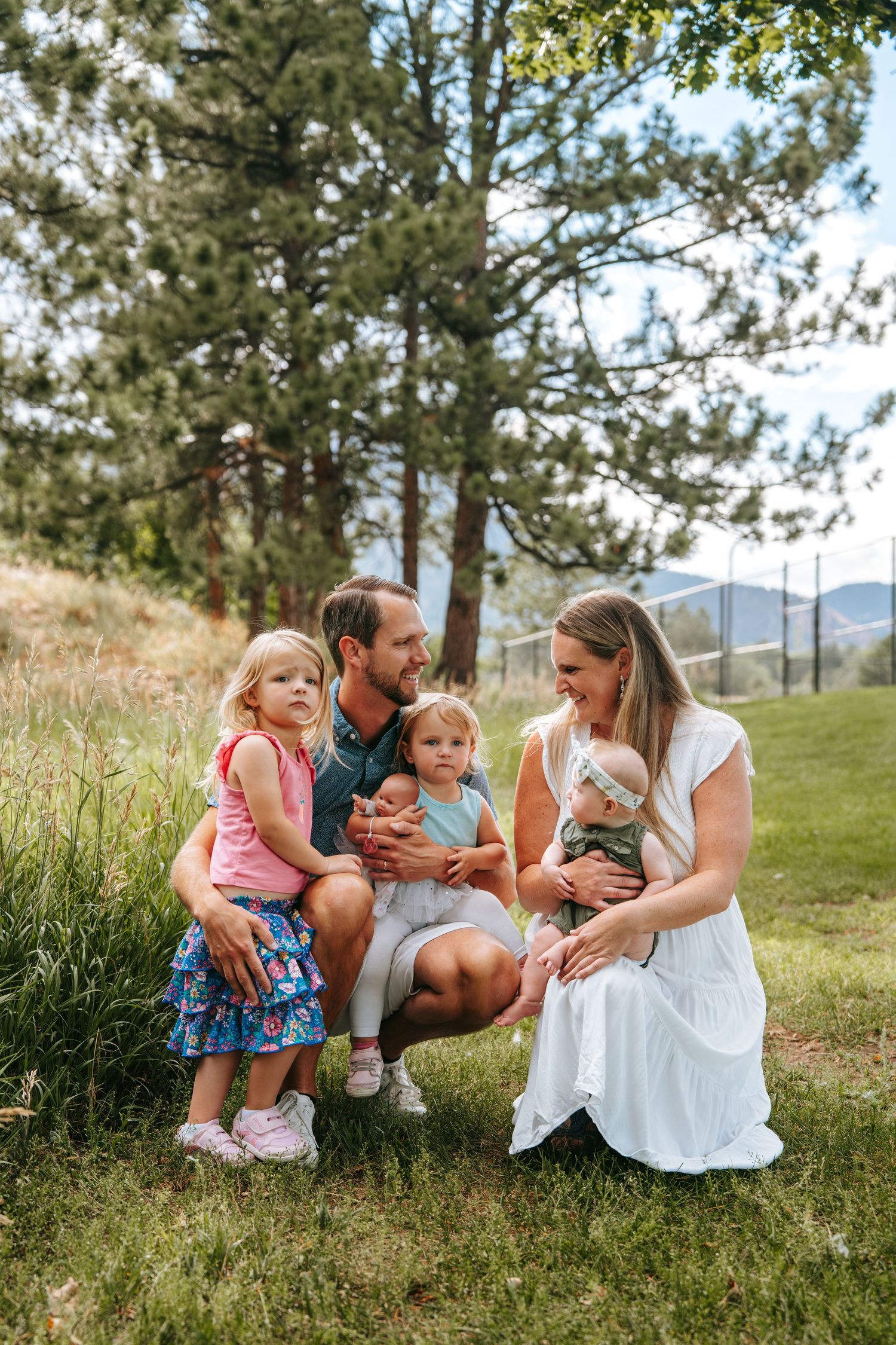 Family of five posing outdoors on grass surrounded by pine trees and mountain views on a sunny day.