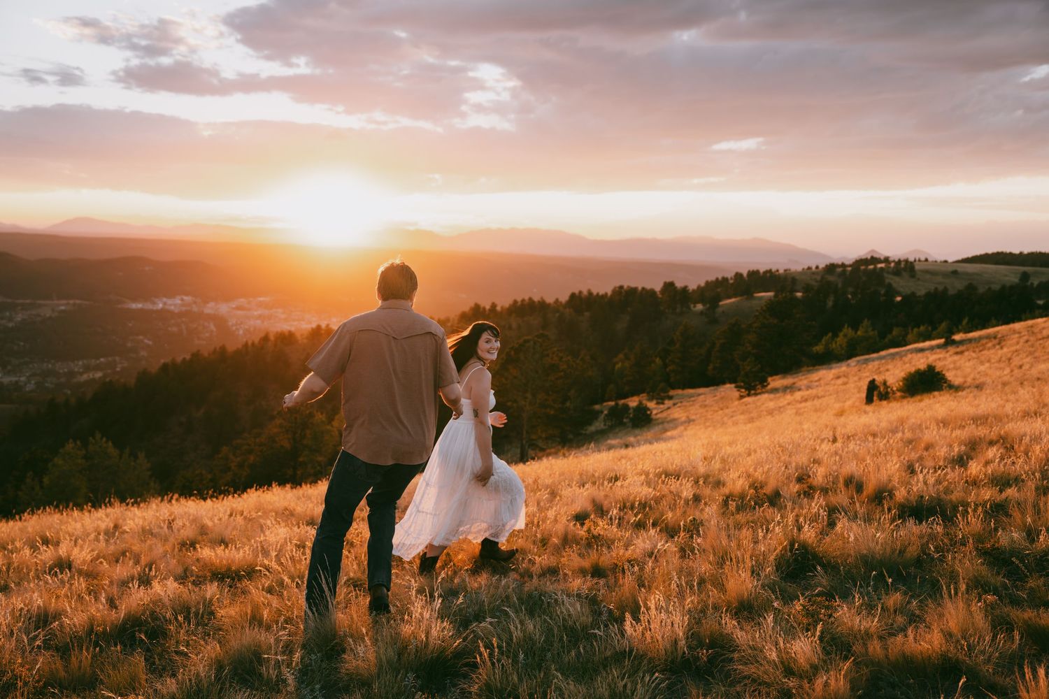 Couple holding hands running through golden meadow at sunset with mountain valley views in romantic outdoor setting.