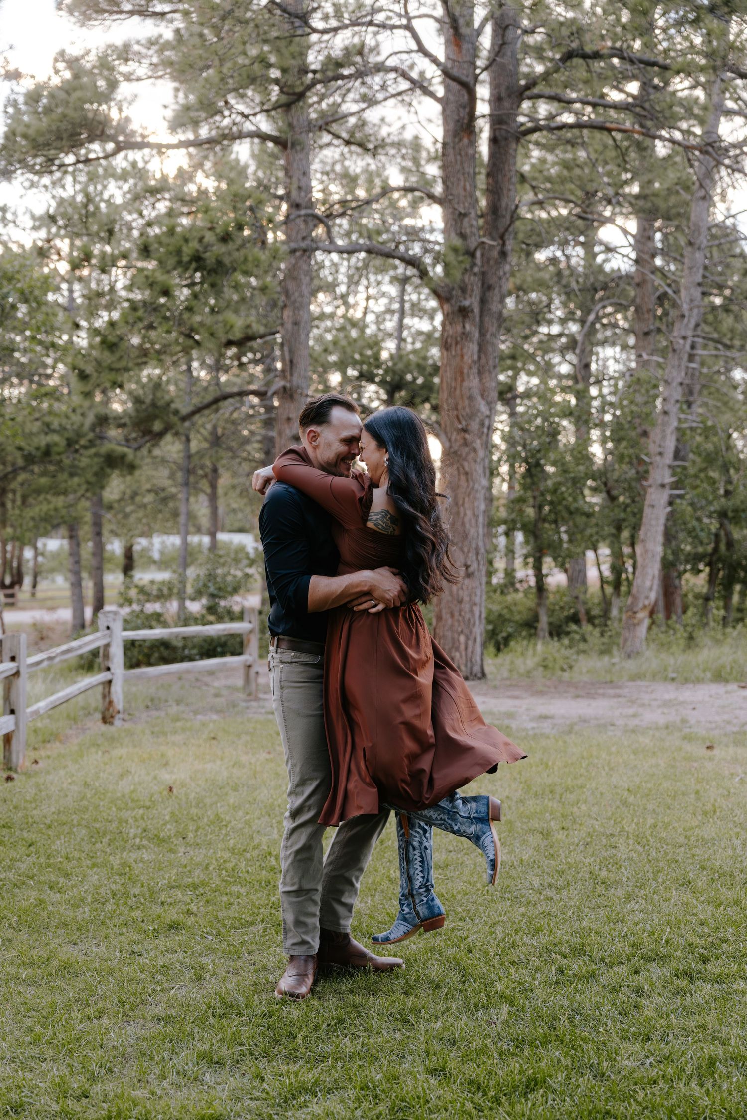 A couple embraces and kisses outdoors on green grass surrounded by tall pine trees in a romantic setting.