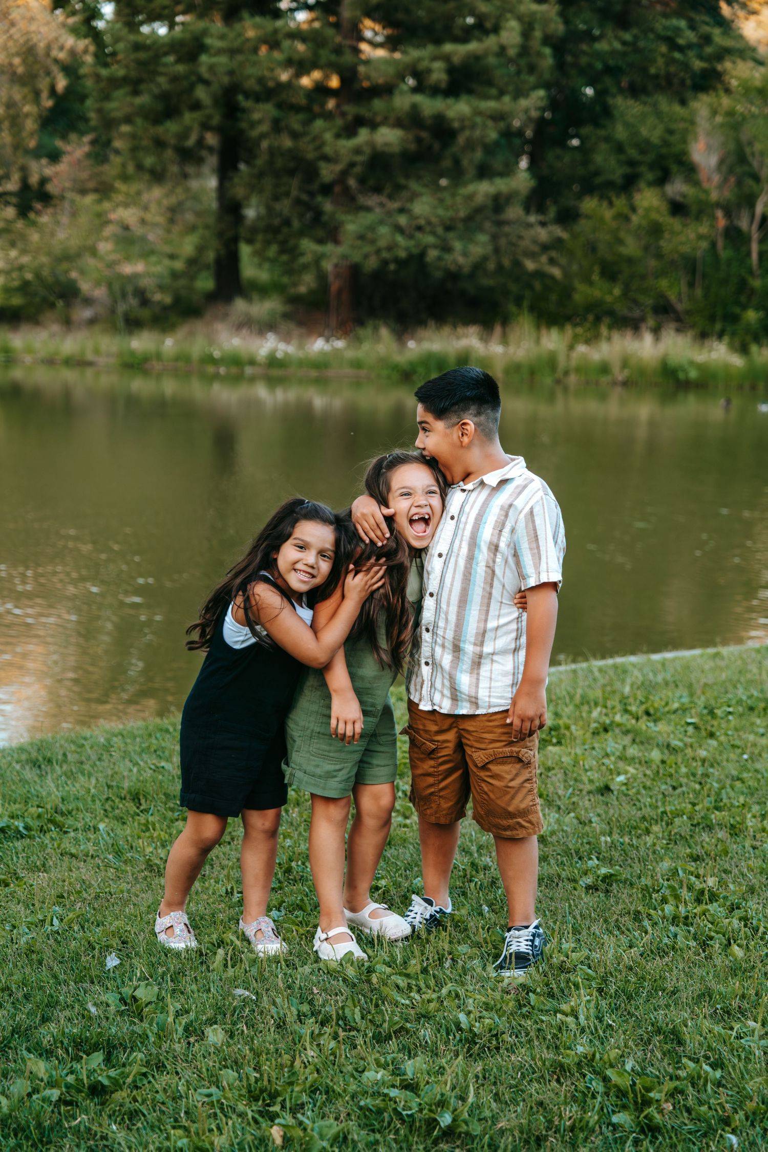 Three siblings hugging and laughing together on green grass beside a tranquil pond in a park.