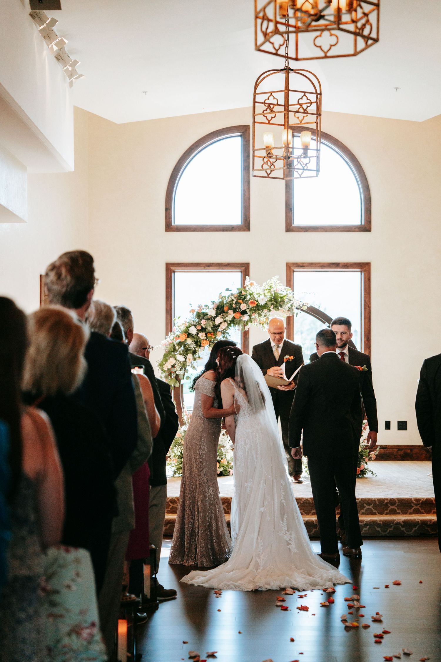 Bride walks down the aisle in a lace gown past seated guests under ornate chandeliers in bright chapel.