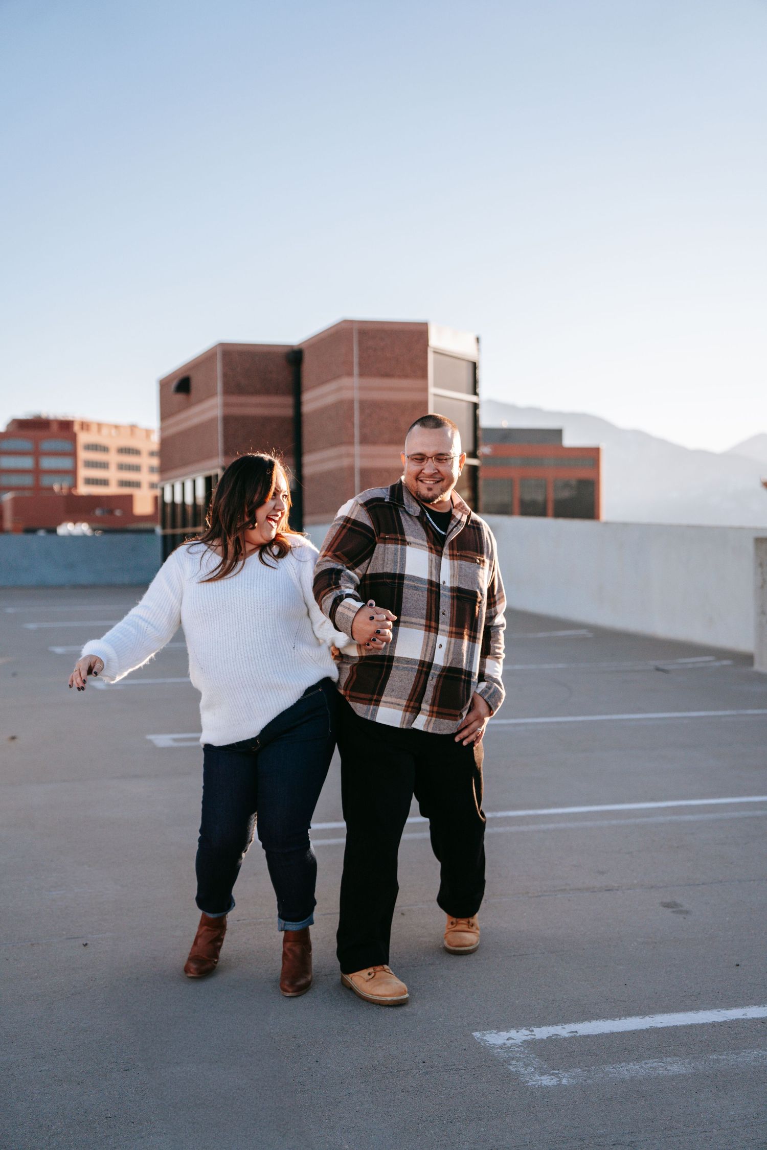 Laughing couple holding hands on a sunny rooftop parking lot with mountains visible in the distance.