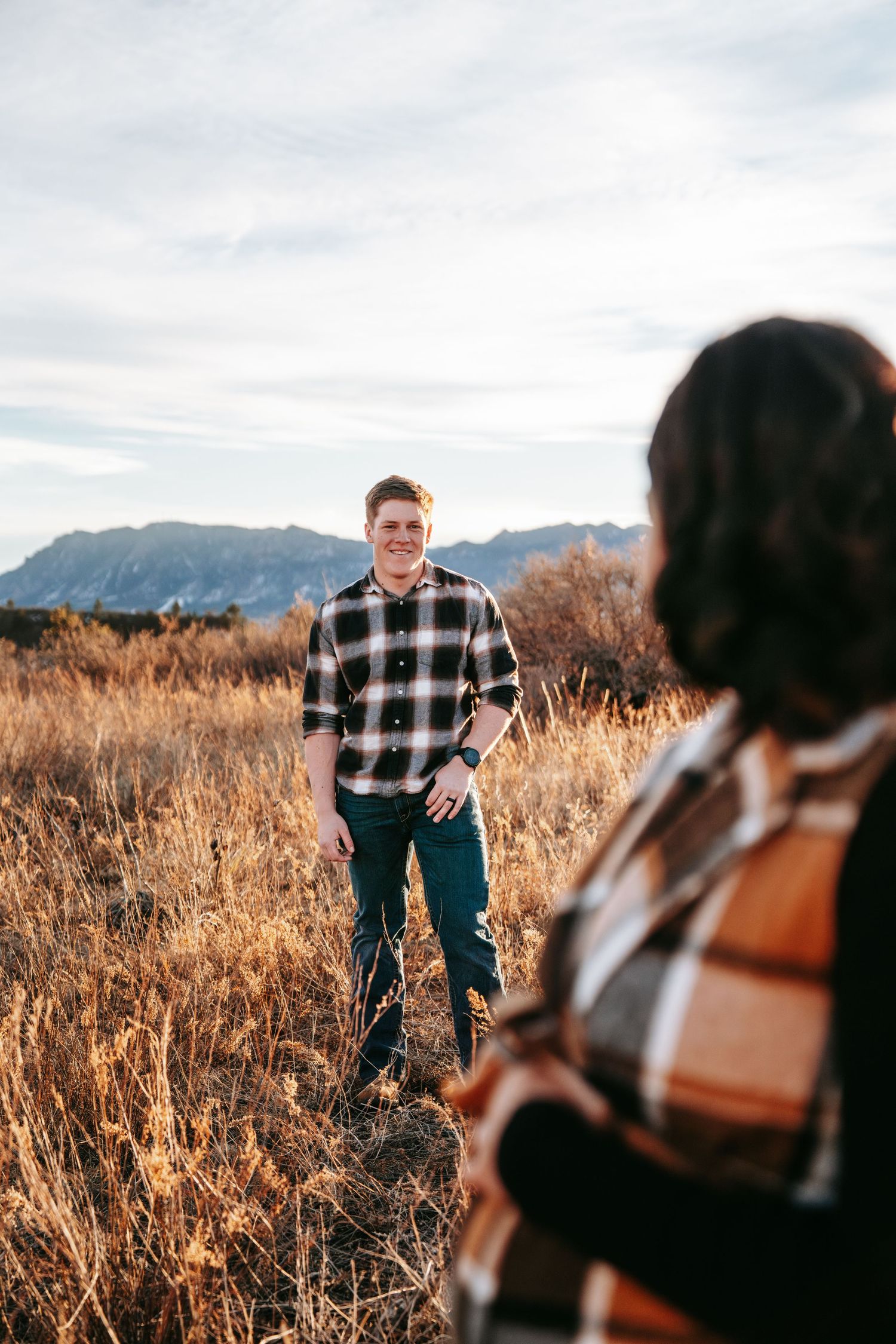 Smiling man in plaid shirt standing in golden field with mountains while pregnant partner stands nearby.